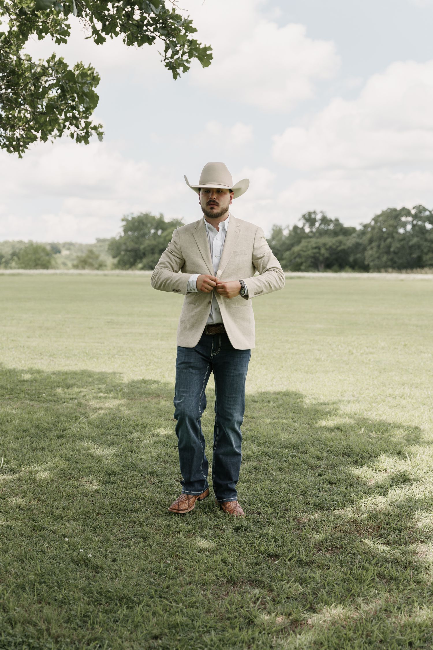 Groom in a cream cowboy hat, tan blazer, jeans and brown boots buttons his jacket while standing on a sunny grassy field.