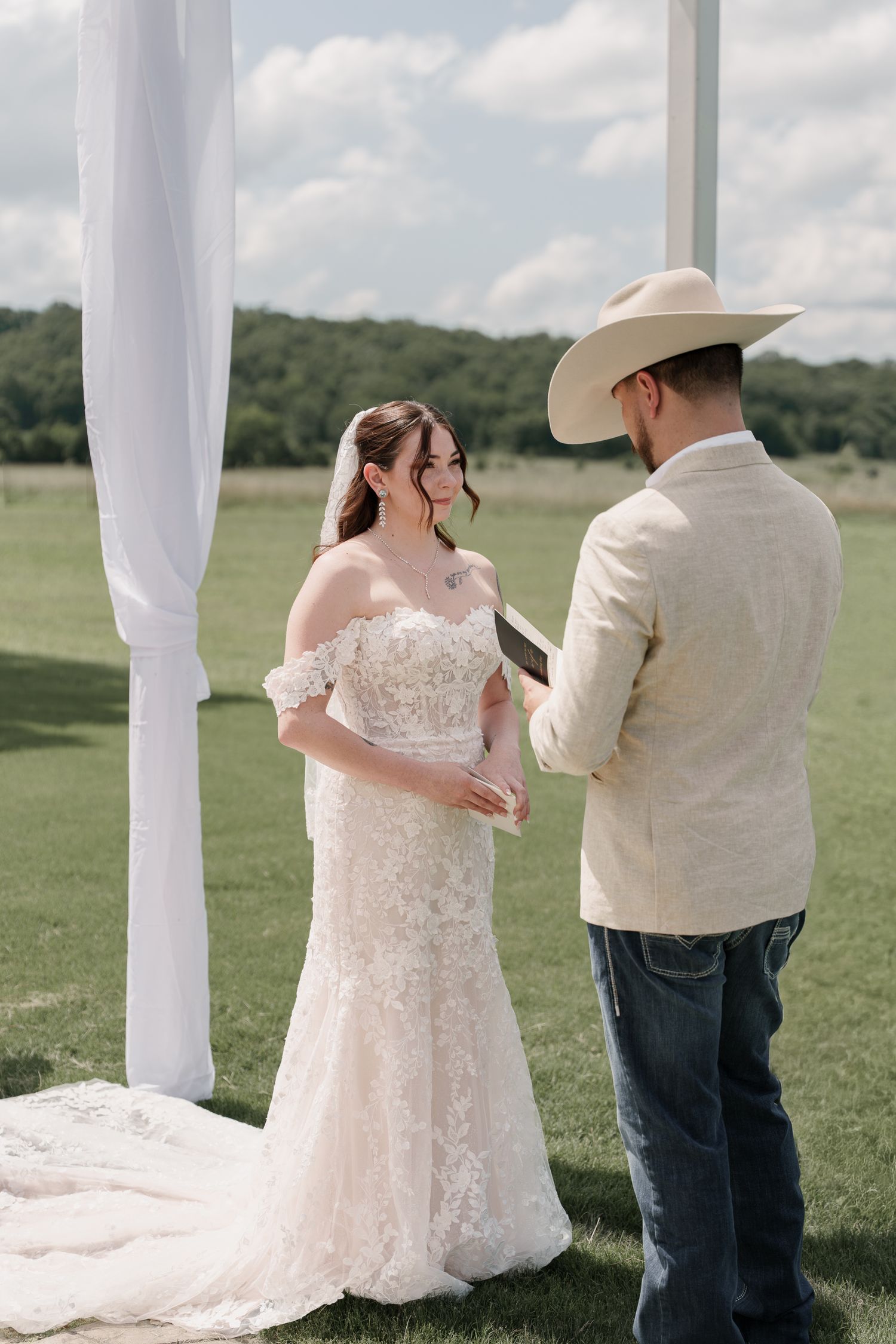 Bride in a lace mermaid wedding gown listens as the groom in a cowboy hat reads his vows during an outdoor ceremony.