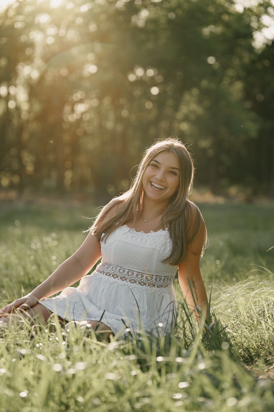 Young woman in a white sundress sits laughing in tall grass as warm golden sunlight glows through the trees behind her.