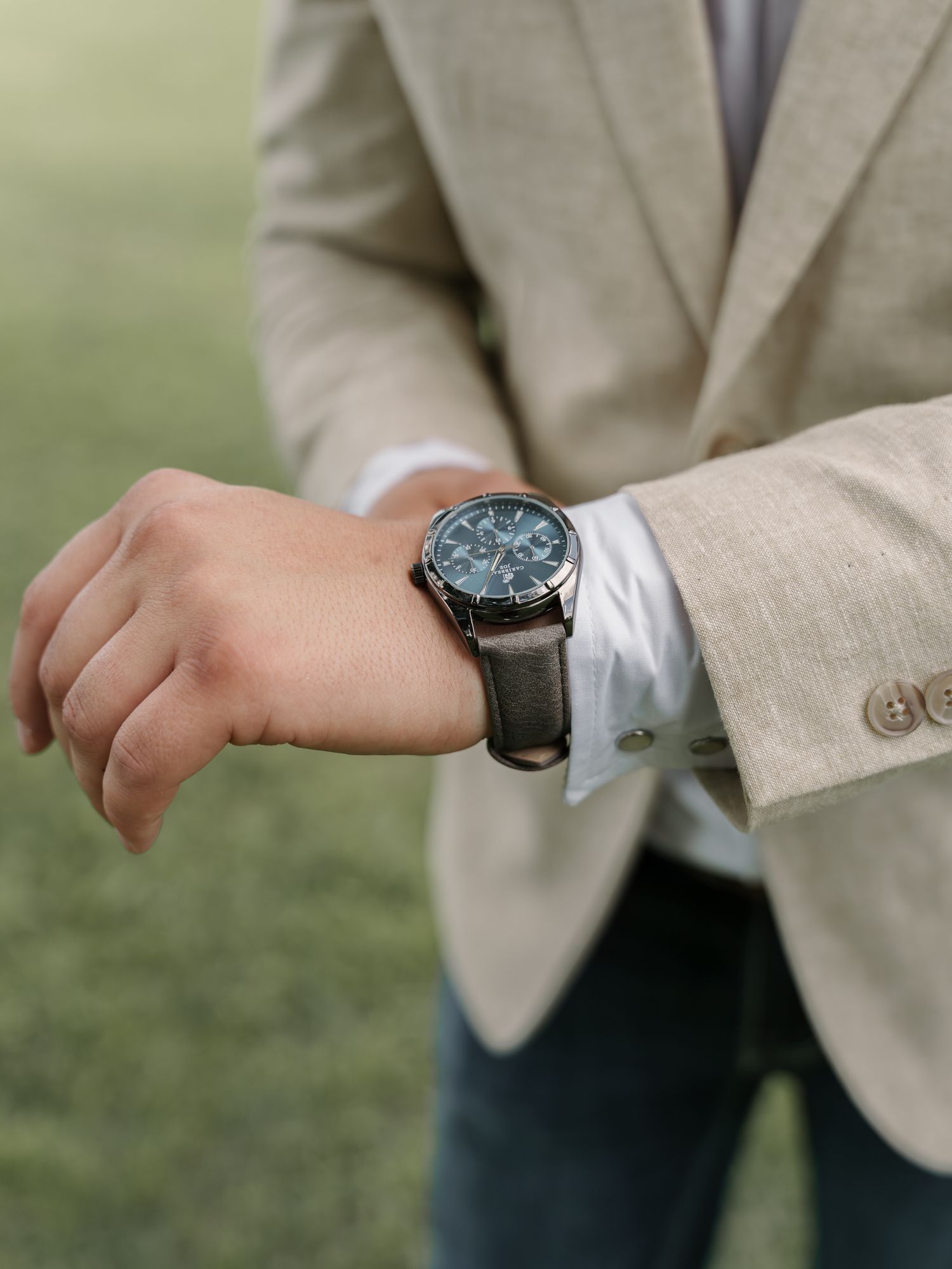 Groom in a tan linen blazer and jeans checks the leather-strap wristwatch on his wrist while standing on a grassy lawn.