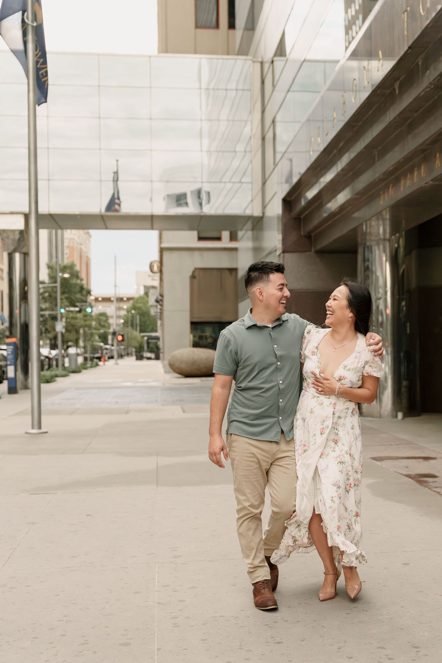 A smiling couple in casual summer outfits walk arm in arm along a city sidewalk past modern glass buildings.