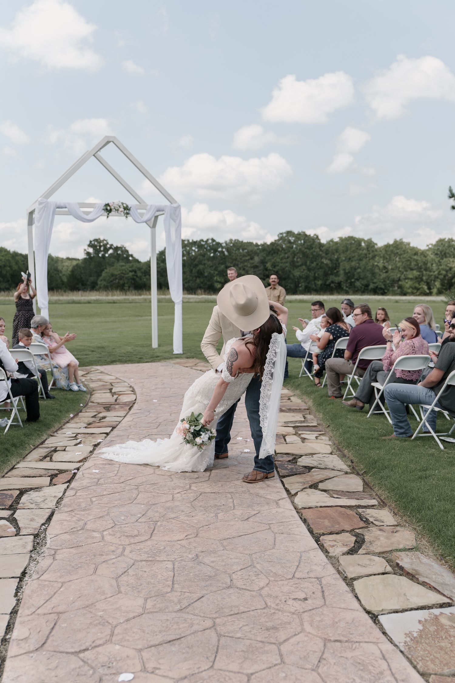 Groom in a cowboy hat dips his bride in a lace wedding gown on a stone aisle as guests cheer at an outdoor ceremony.