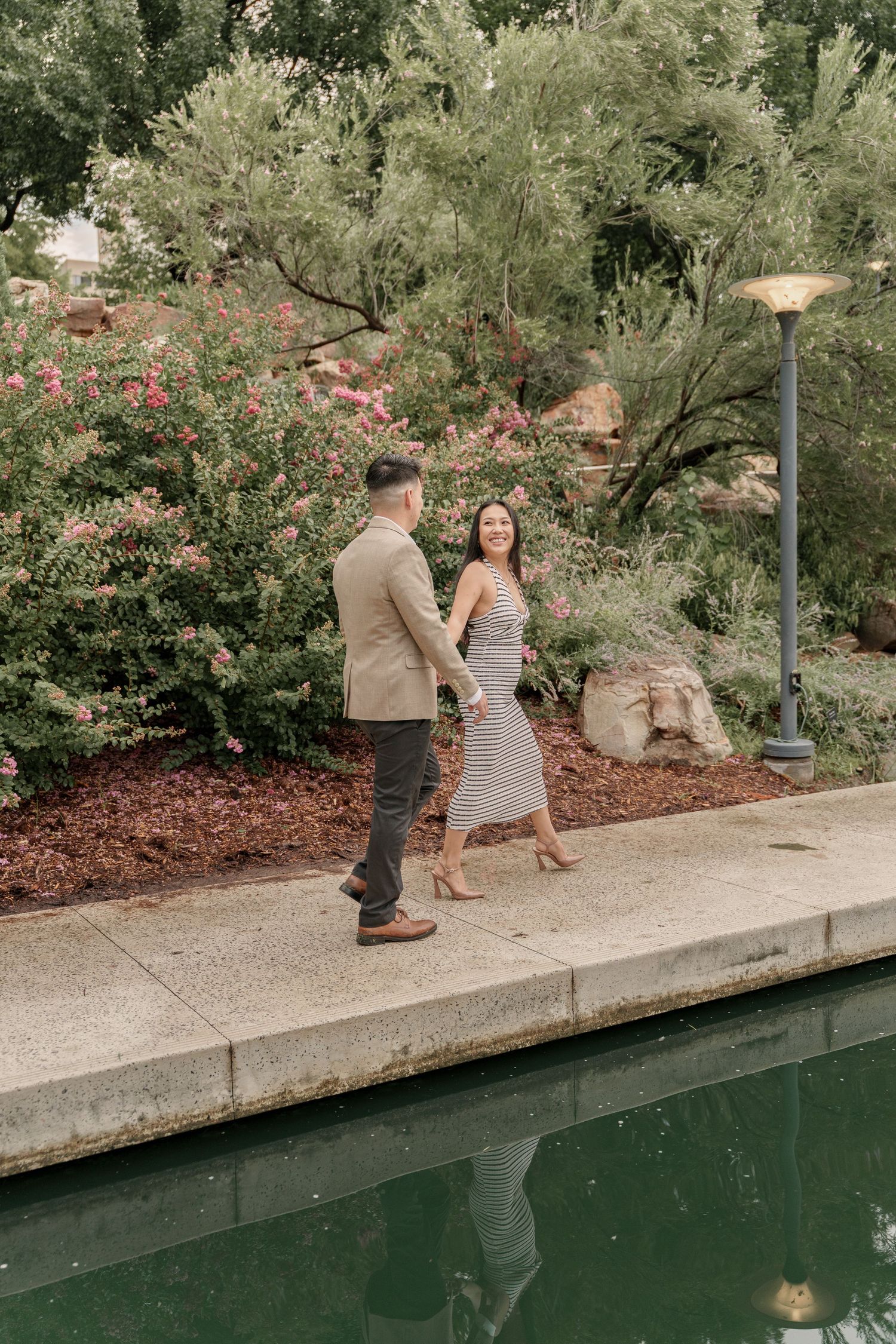 An engaged couple walk hand in hand along a stone path beside a reflecting pool and blooming pink shrubs.
