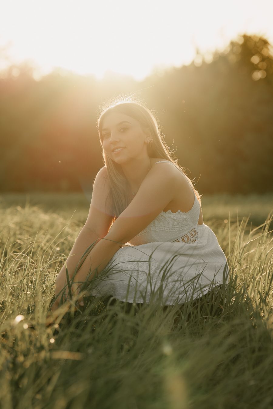Young woman in a white dress crouching in a golden meadow at sunset with warm sunlight glowing behind her.