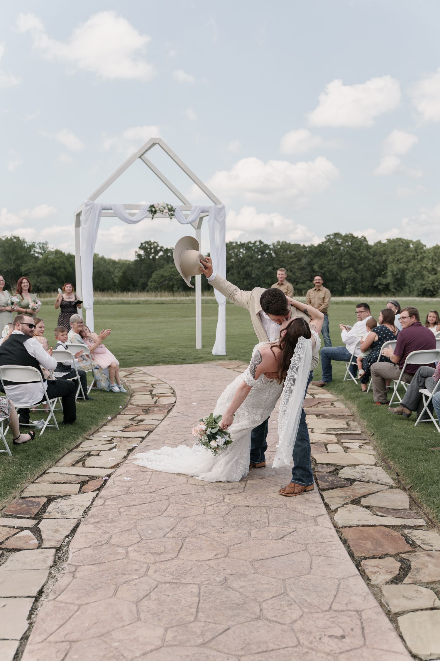 Groom dips the bride in a romantic kiss on a stone aisle near a white wedding arch as guests look on.