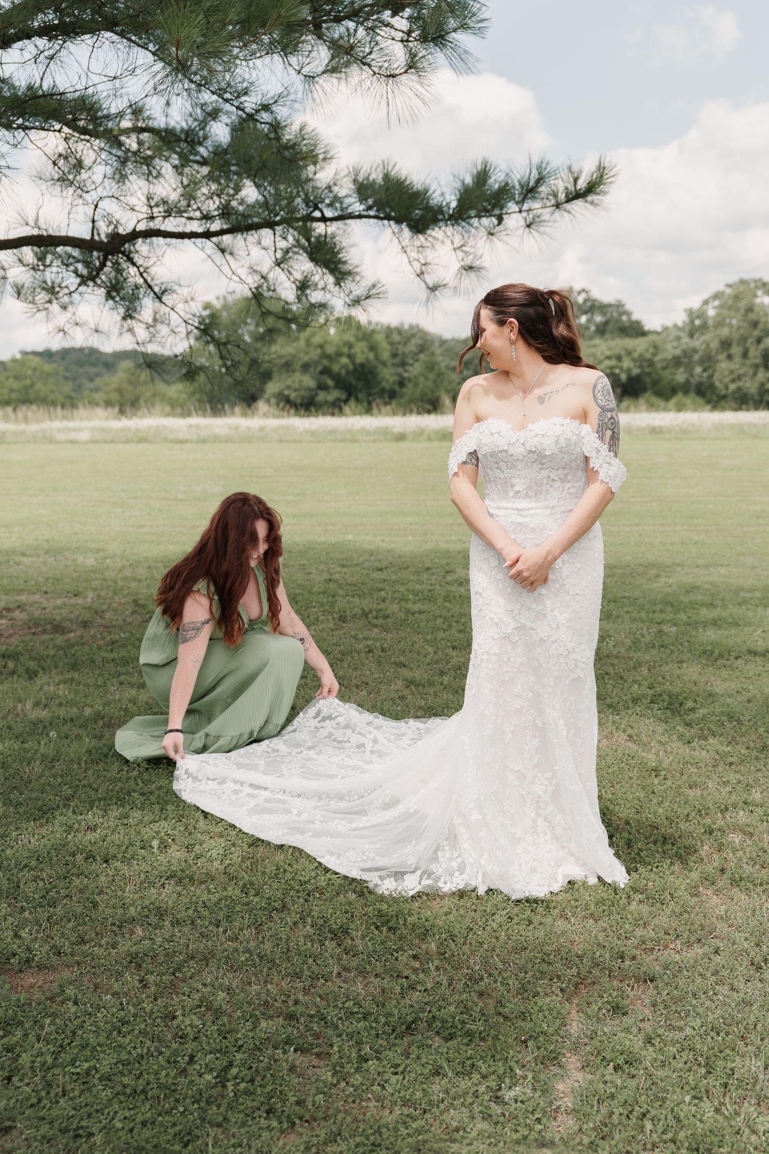 Bridesmaid in a green dress kneels to arrange the train of a bride's lace wedding gown on a grassy lawn under a pine tree.