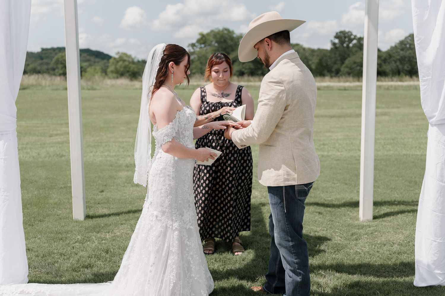 Bride and groom in a cowboy hat exchange vows with the officiant under a white wedding arch in a green field.