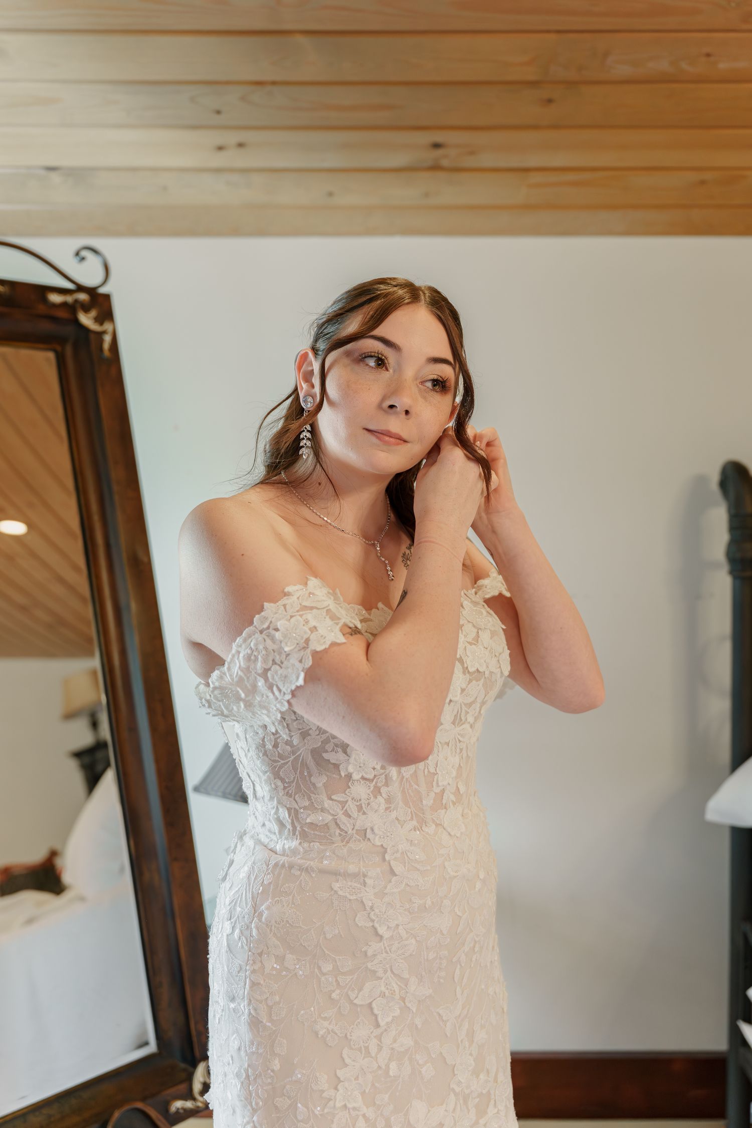 Bride in an off-shoulder lace wedding gown adjusts her earring in front of a mirror in a rustic room with wood ceiling.