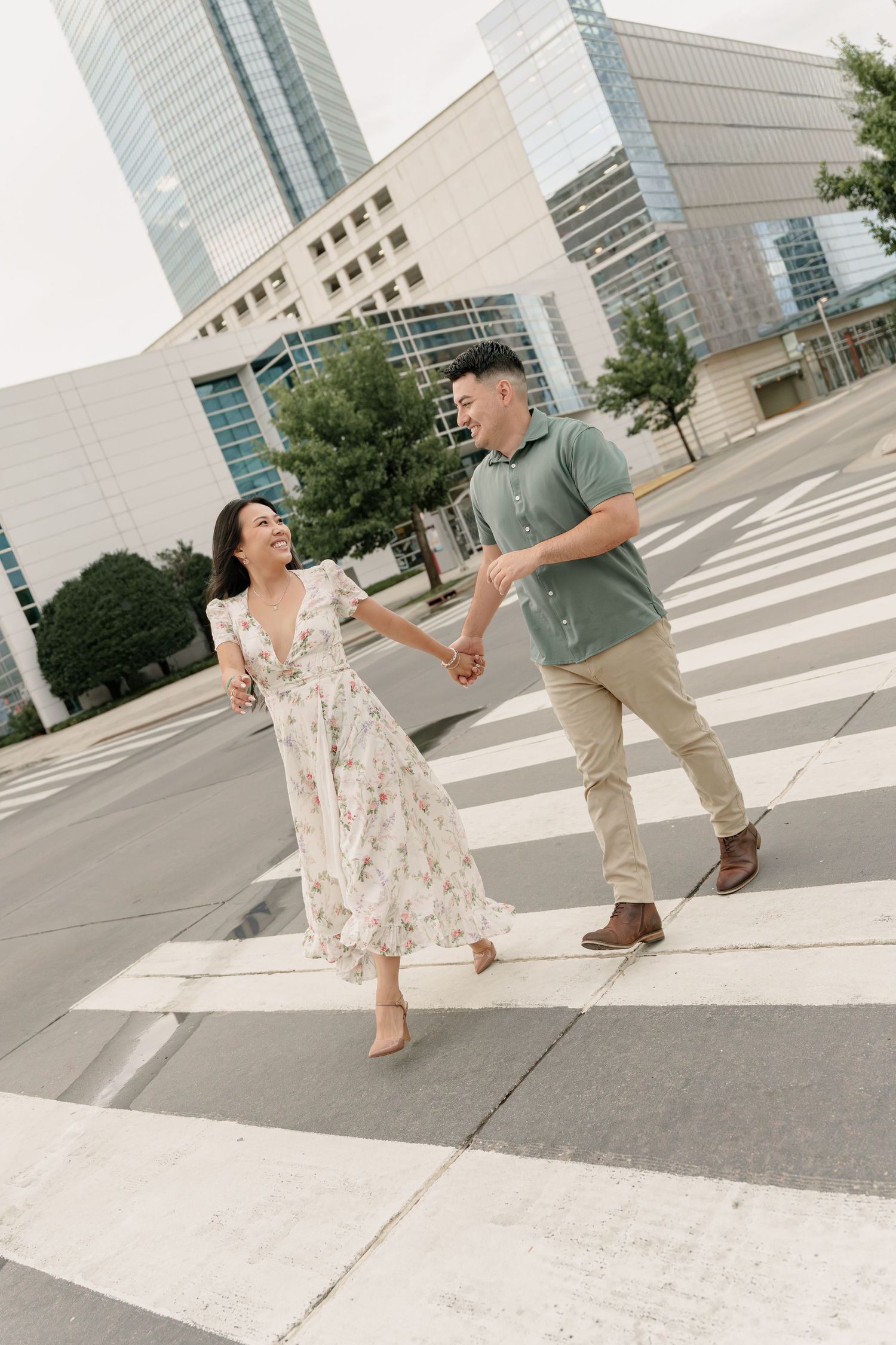 Happy couple holds hands and runs across a downtown crosswalk, she in a floral dress and he in a green shirt, for engagement photos.