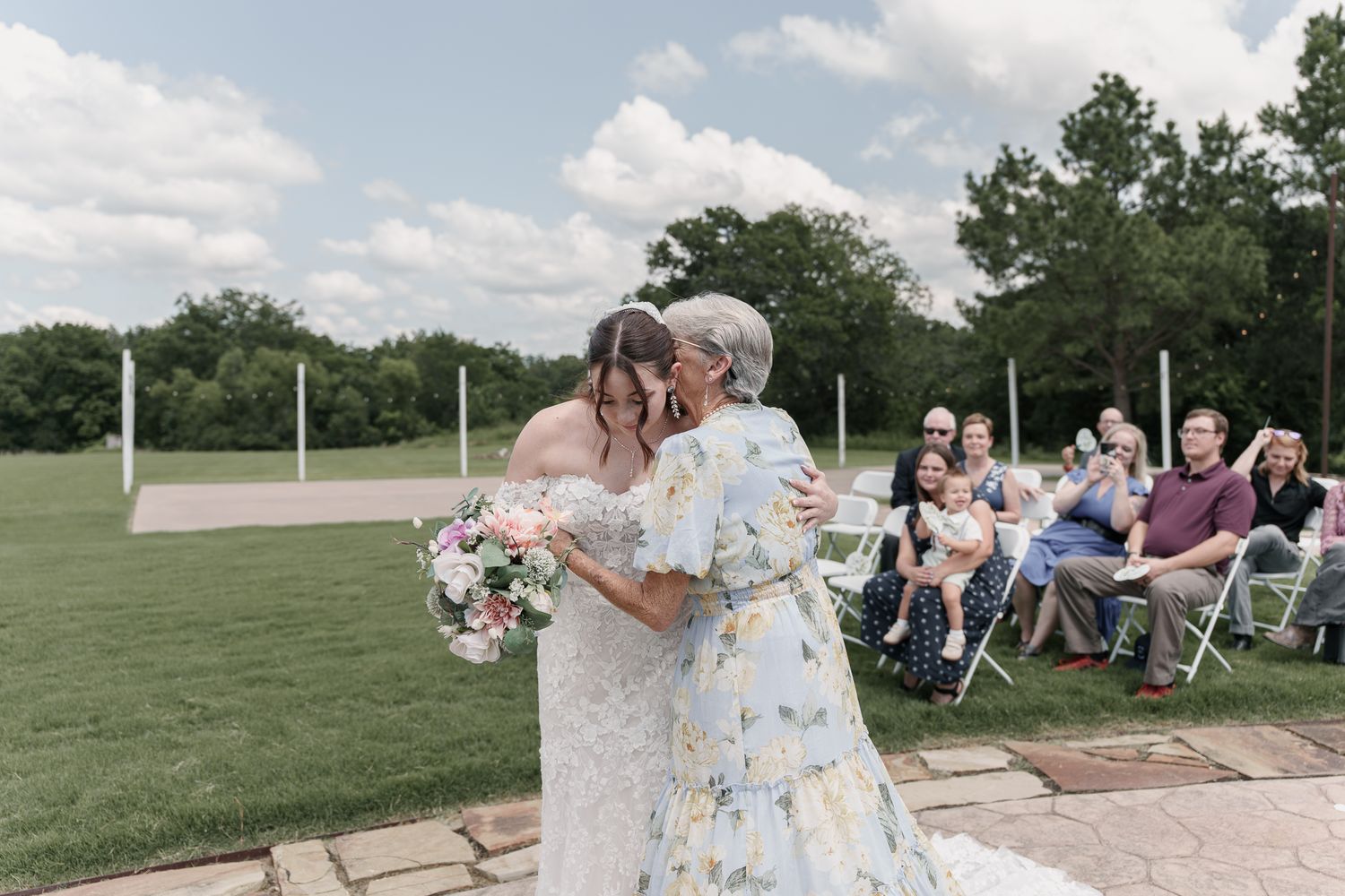 Bride in a lace wedding dress embraces a woman in a floral dress as guests watch from white chairs outdoors.