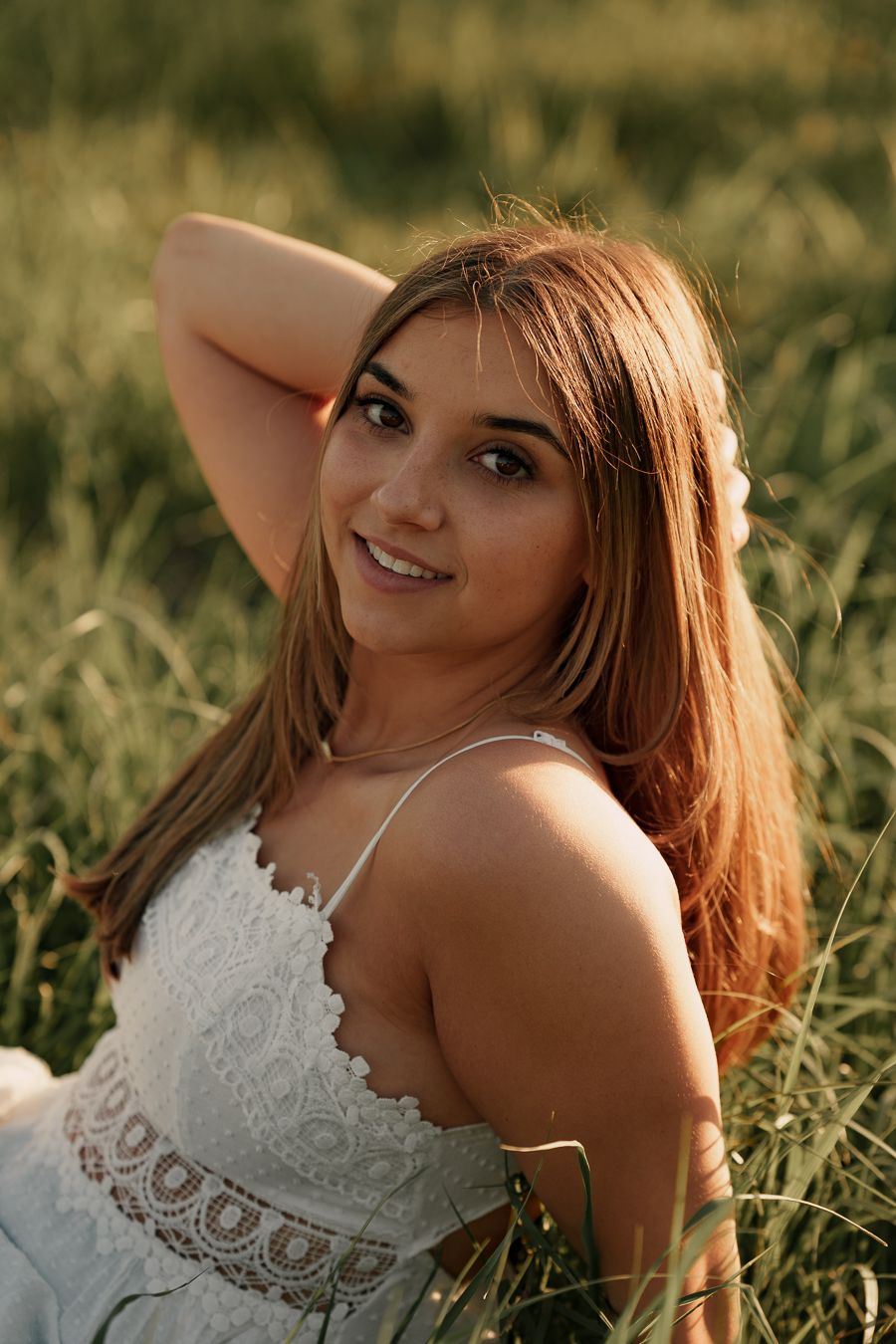 Smiling young woman with long hair in a white lace sundress resting in tall green grass with her arm raised behind her head.