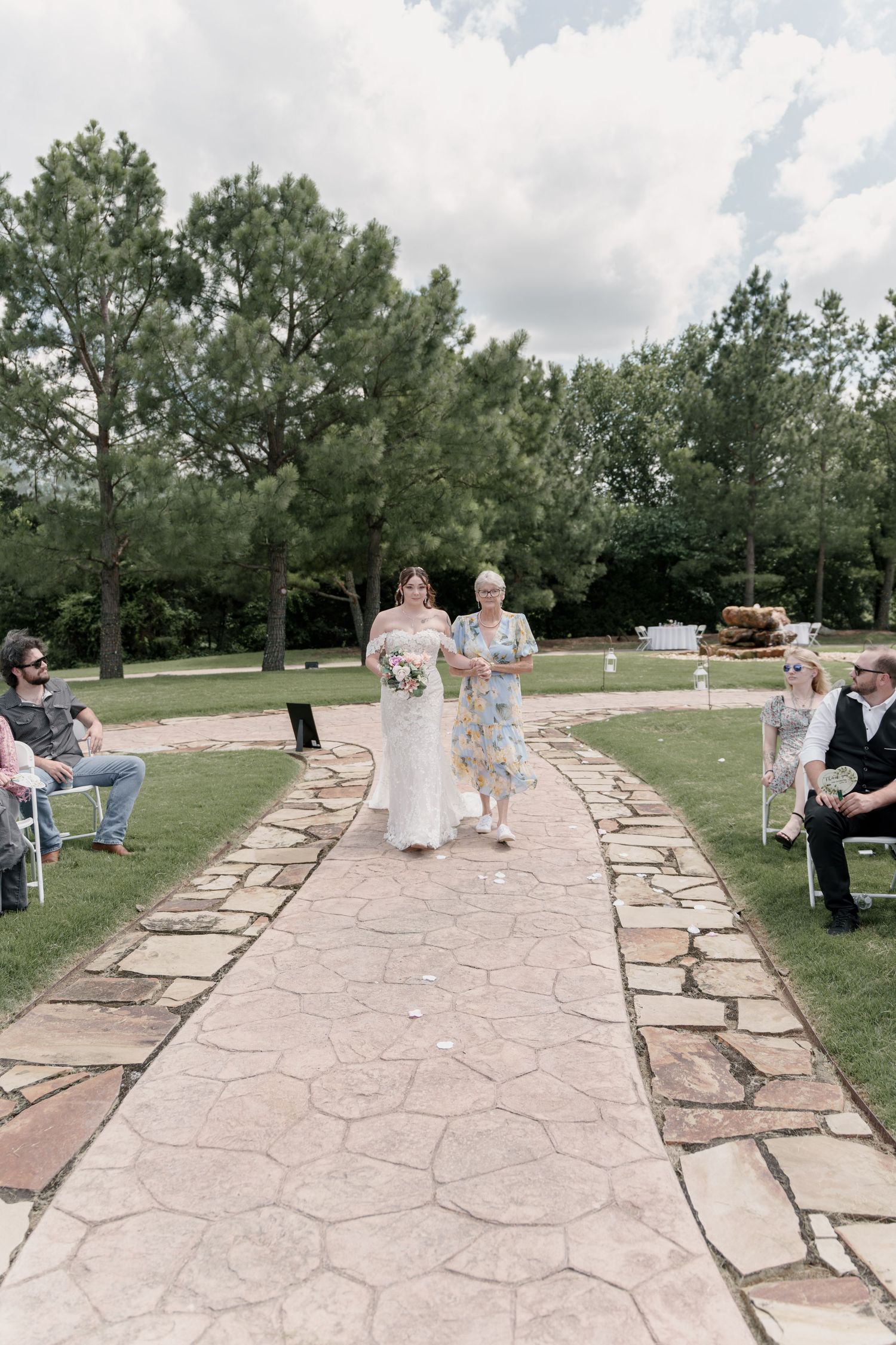 Bride in a white gown walks down a stone aisle with an older woman as seated wedding guests look on beneath tall green trees.