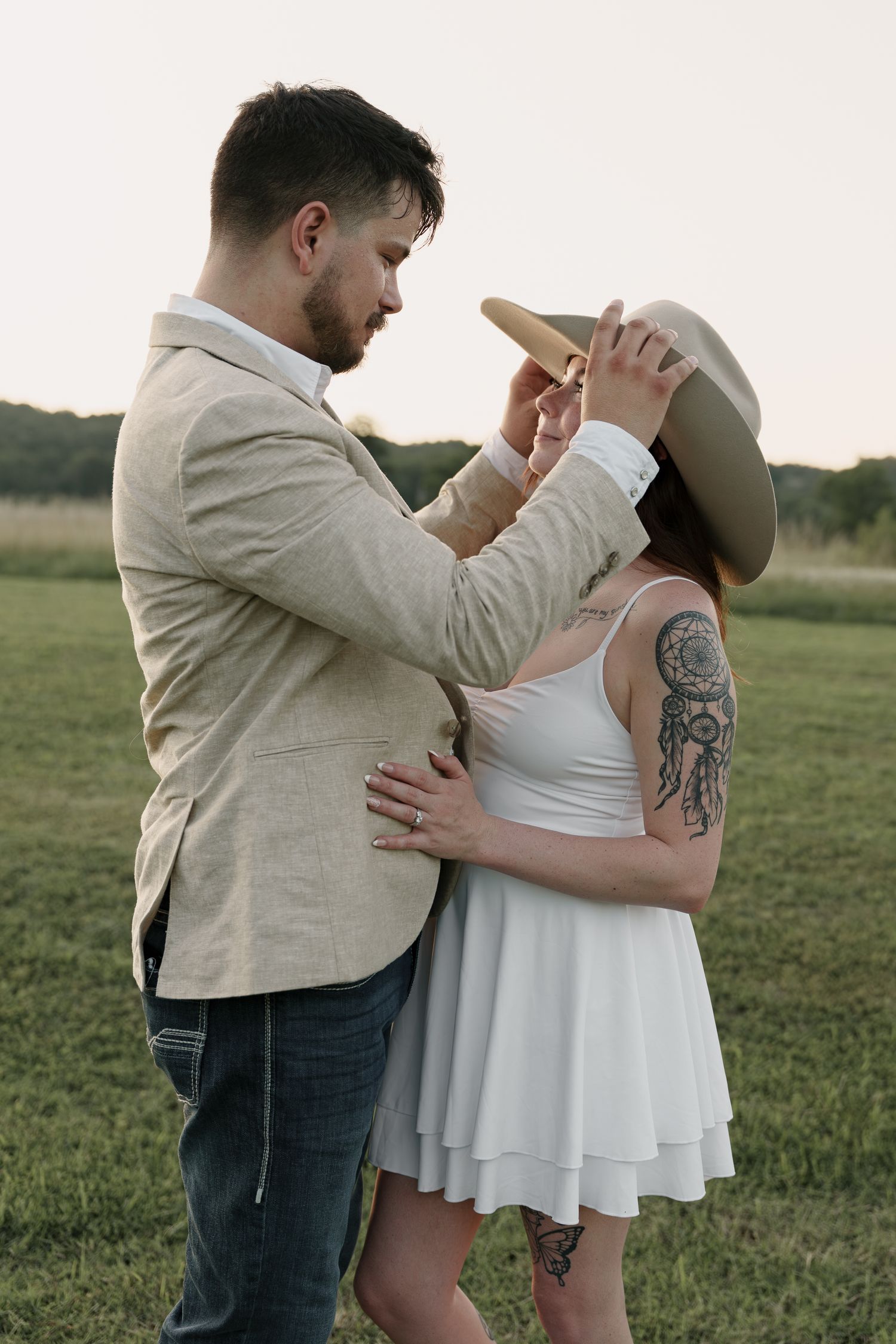 Groom in a linen blazer adjusts a wide-brim hat on his tattooed partner in a white dress in a field at sunset.