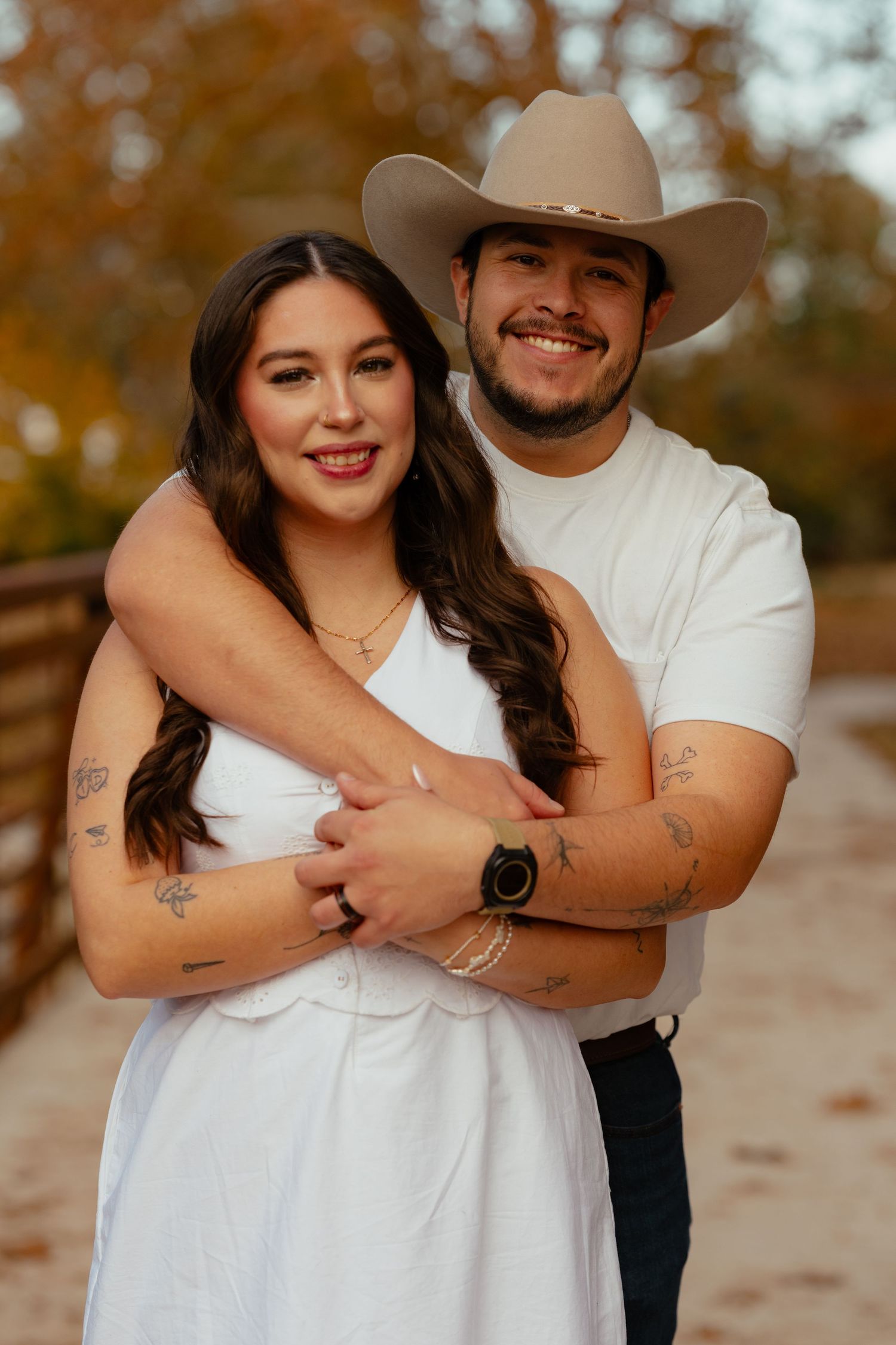 Smiling couple in white outfits embrace outdoors, the man wearing a tan cowboy hat, with autumn trees behind them.