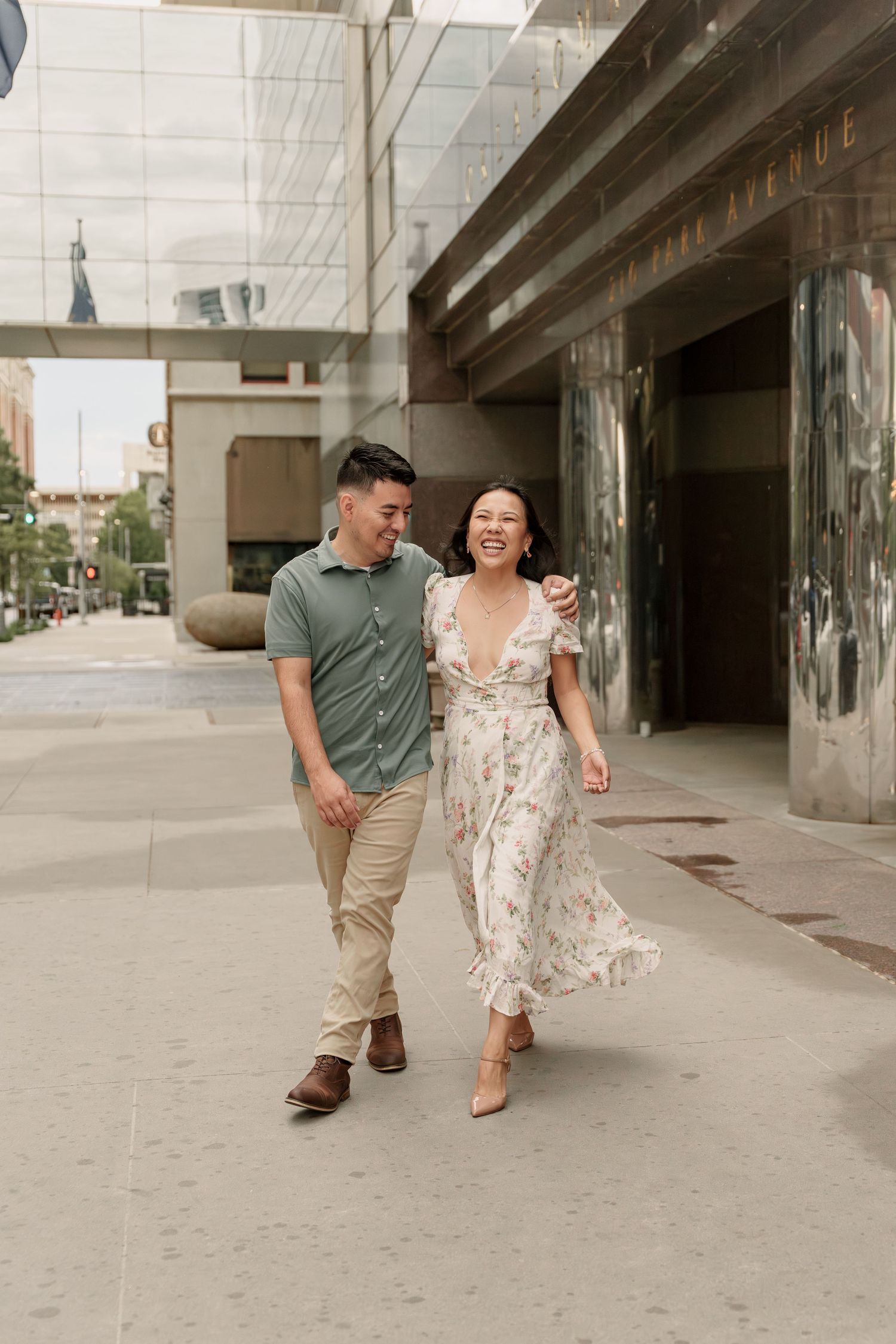 Laughing couple strolls arm in arm along a downtown sidewalk past modern glass and steel buildings in an engagement photo.