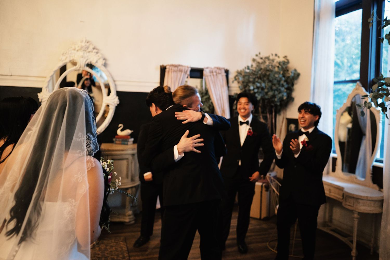 Groom embraces guest warmly while wedding party celebrates inside elegantly decorated wedding reception venue.