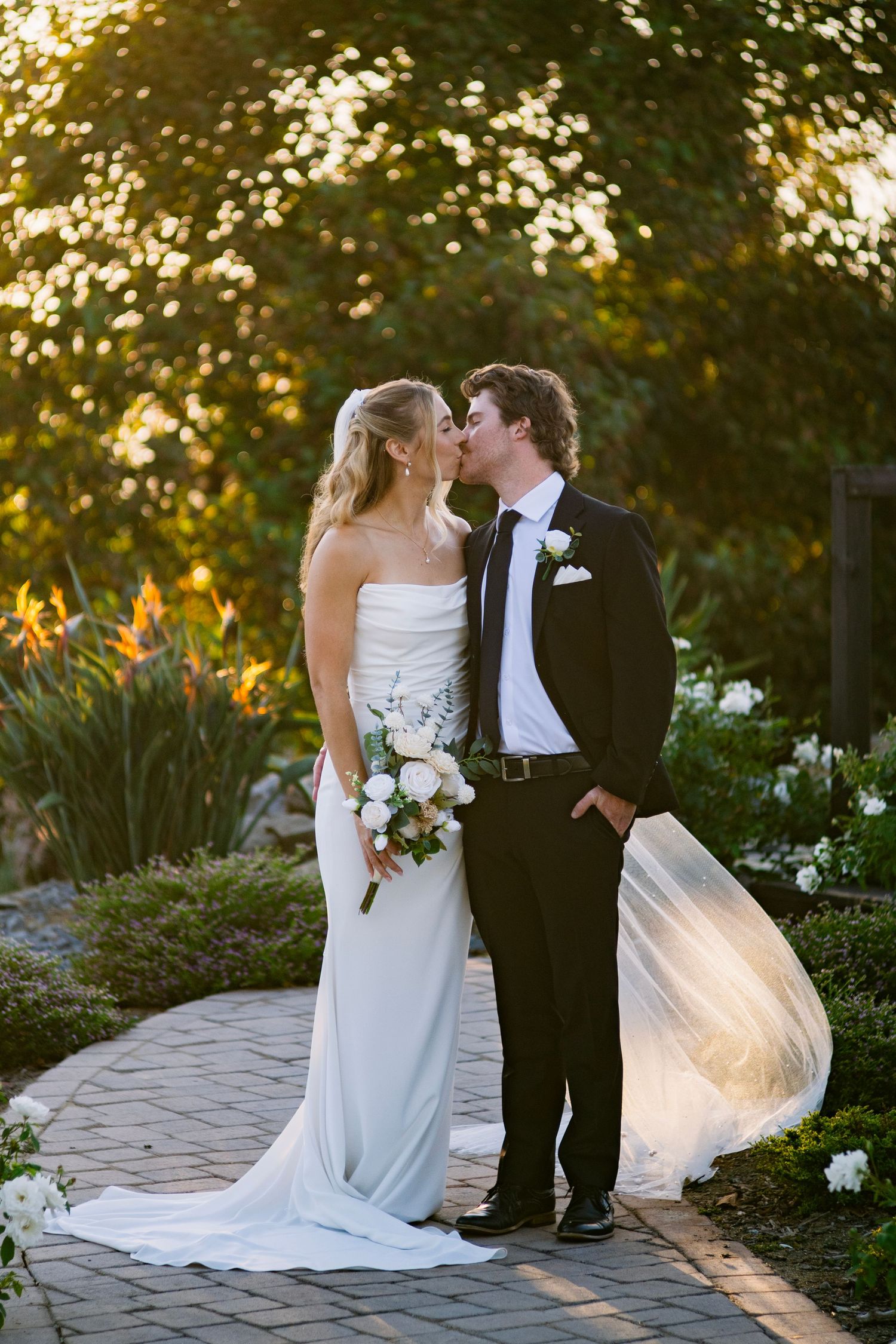 Bride and groom share a romantic kiss on a garden pathway at golden hour, with veil flowing in the breeze.