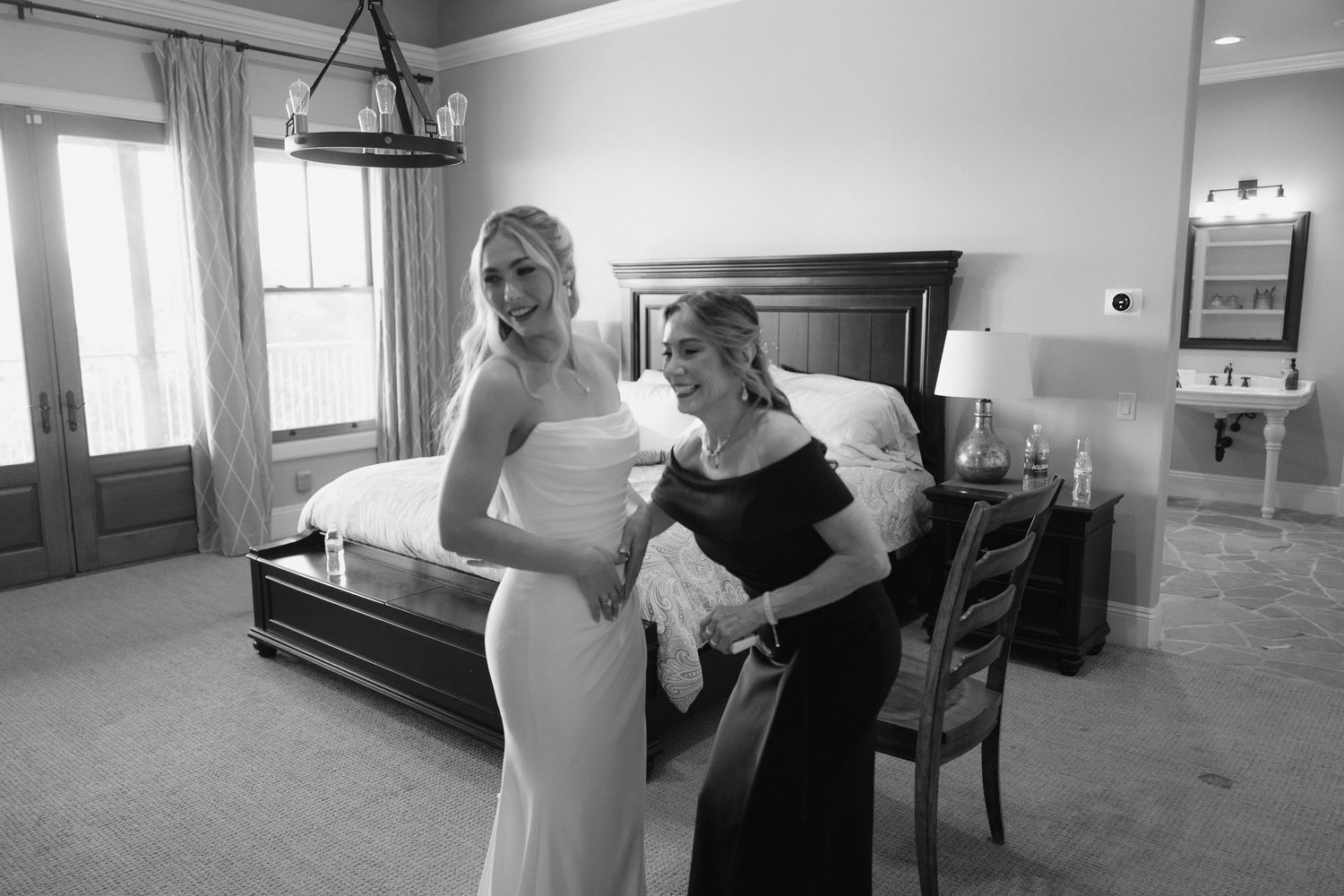 Bride in elegant fitted white gown being helped by mother in black dress, black and white.