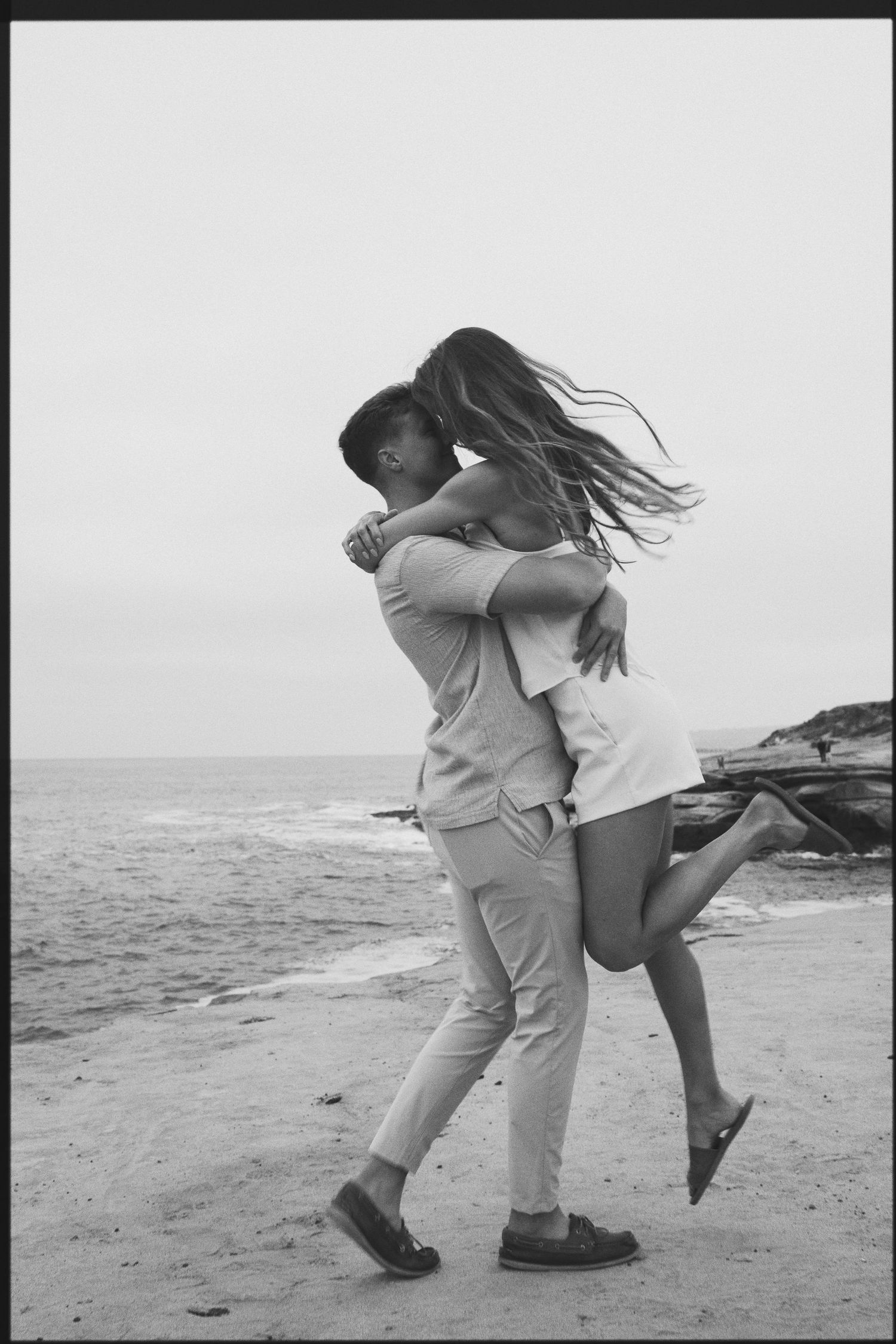 Black and white beach photo of a man joyfully lifting a woman in a romantic embrace.