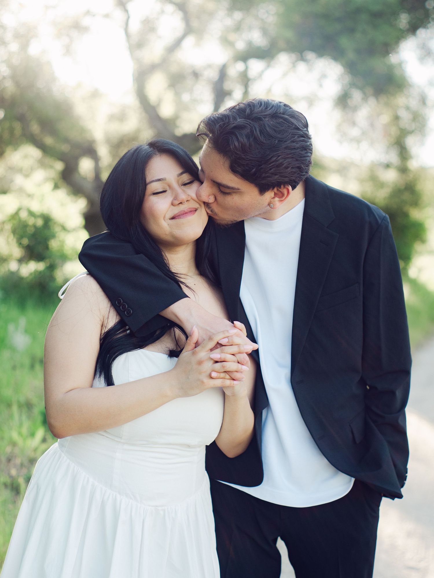 Groom kisses bride on cheek while holding hands on scenic outdoor path during engagement shoot.