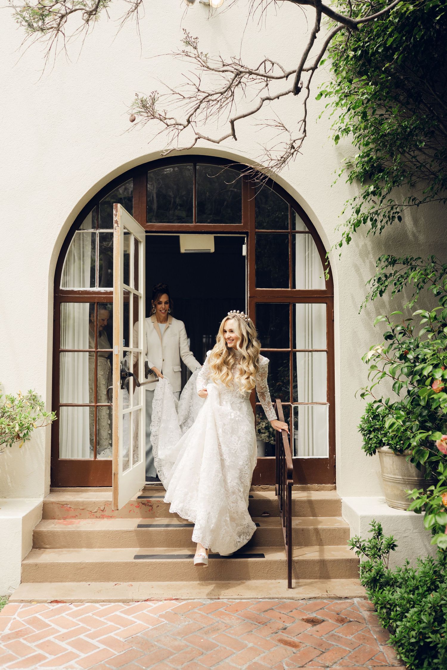 Bride in lace wedding dress descending steps through arched doorway with attendant following behind.