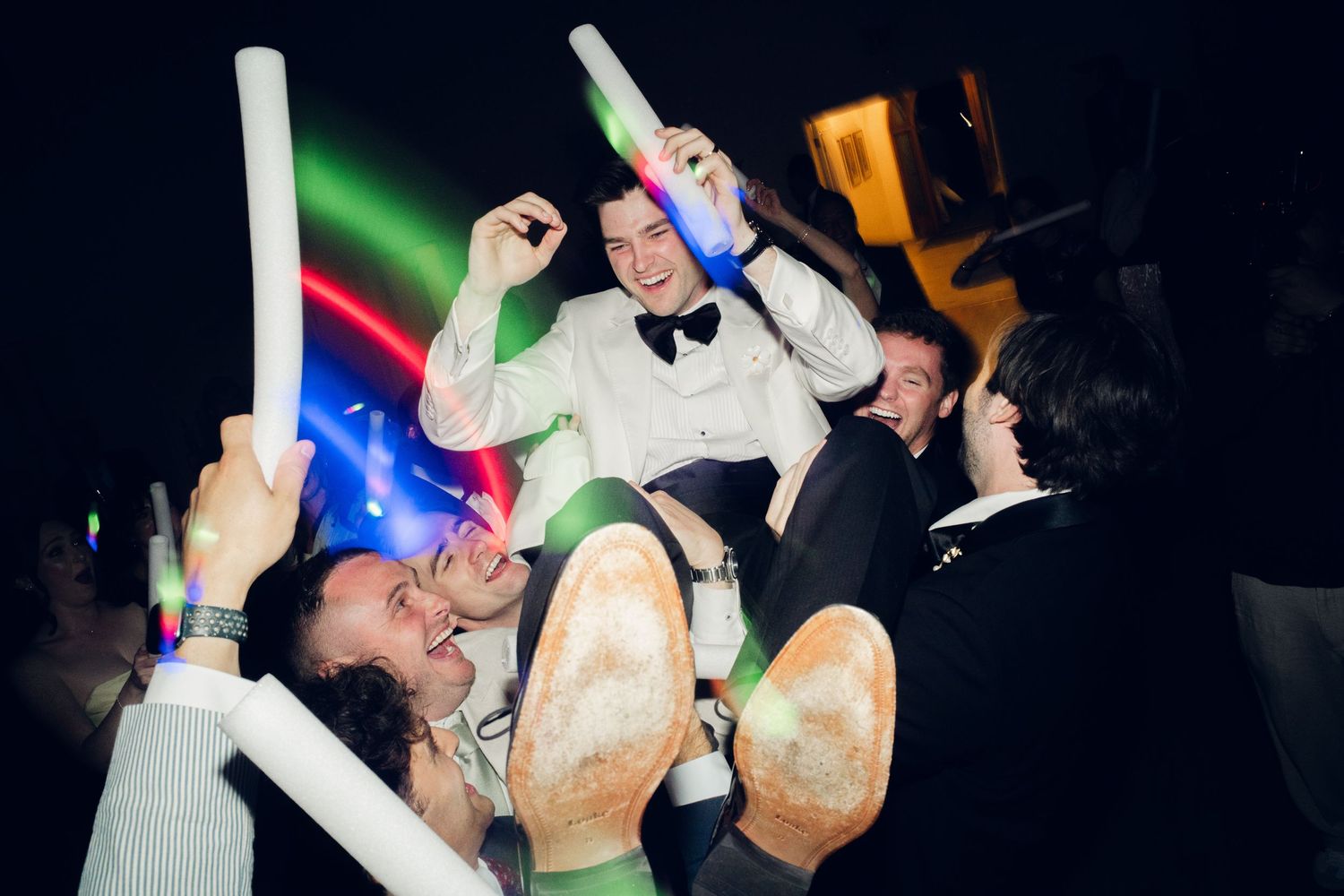 Groom is lifted by cheering guests holding colorful glow sticks at a festive wedding reception.
