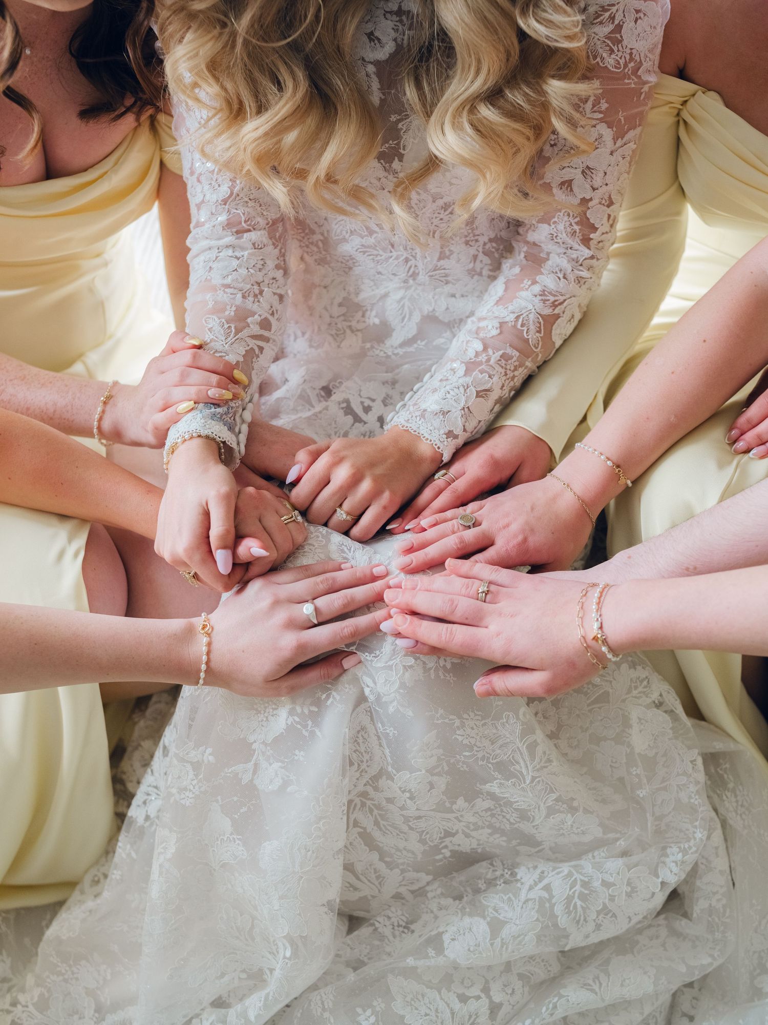 Close-up of bride and bridesmaids showing stacked hands with rings and gold bracelets against white lace wedding gown.