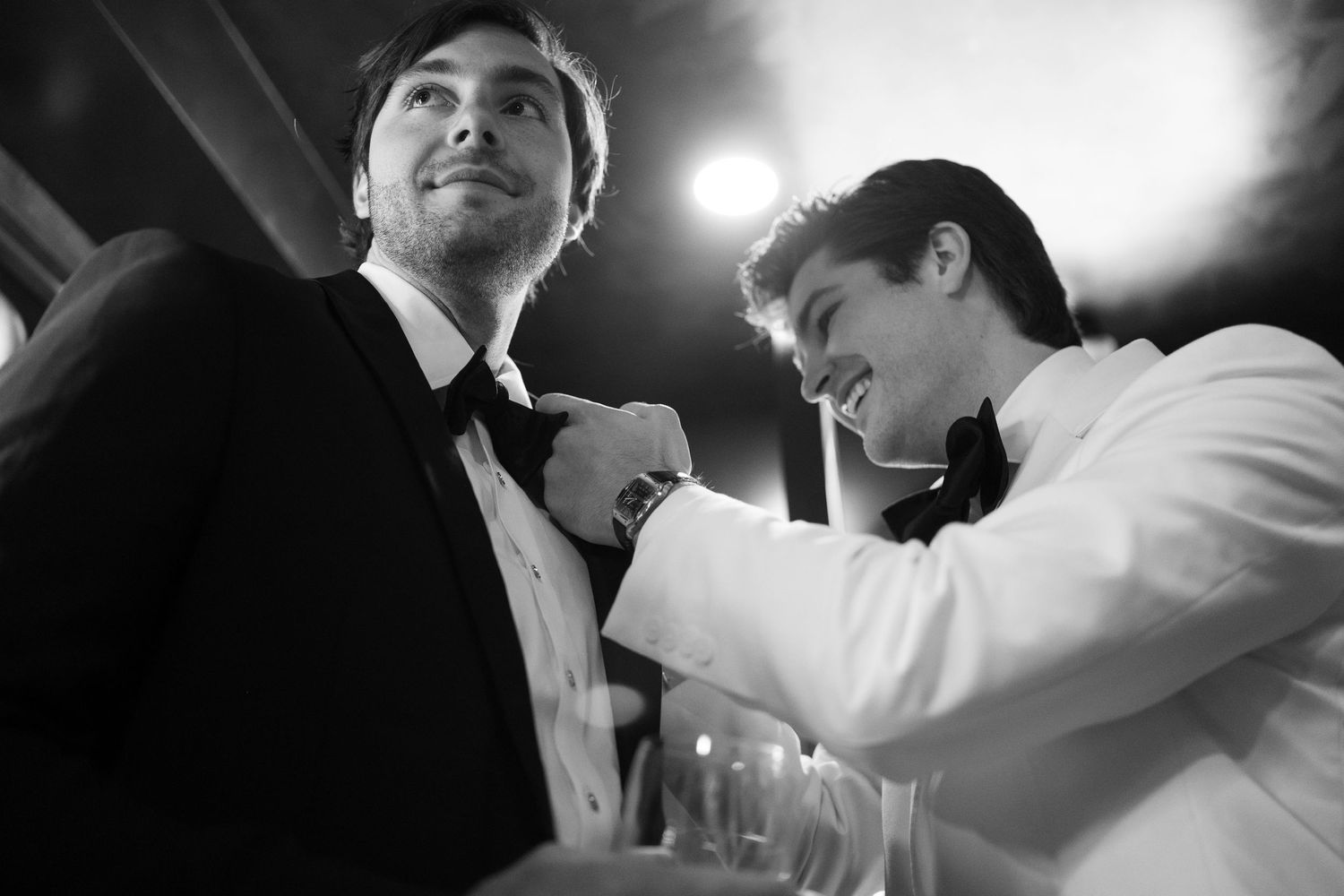 Black and white photo of groom in white jacket adjusting bow tie of groomsman in black tuxedo at wedding.