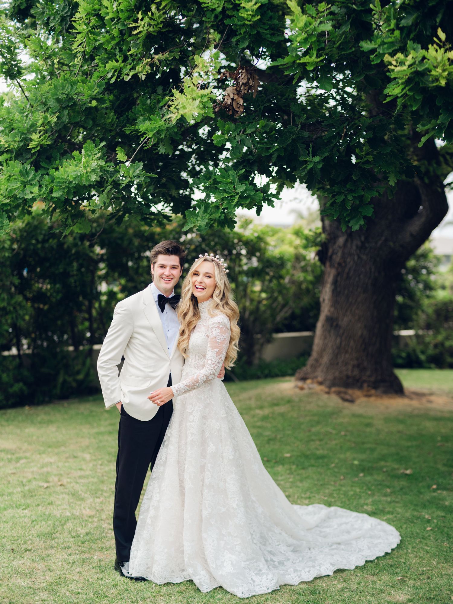 Smiling bride and groom pose together under a large tree, bride wearing lace gown and floral crown.