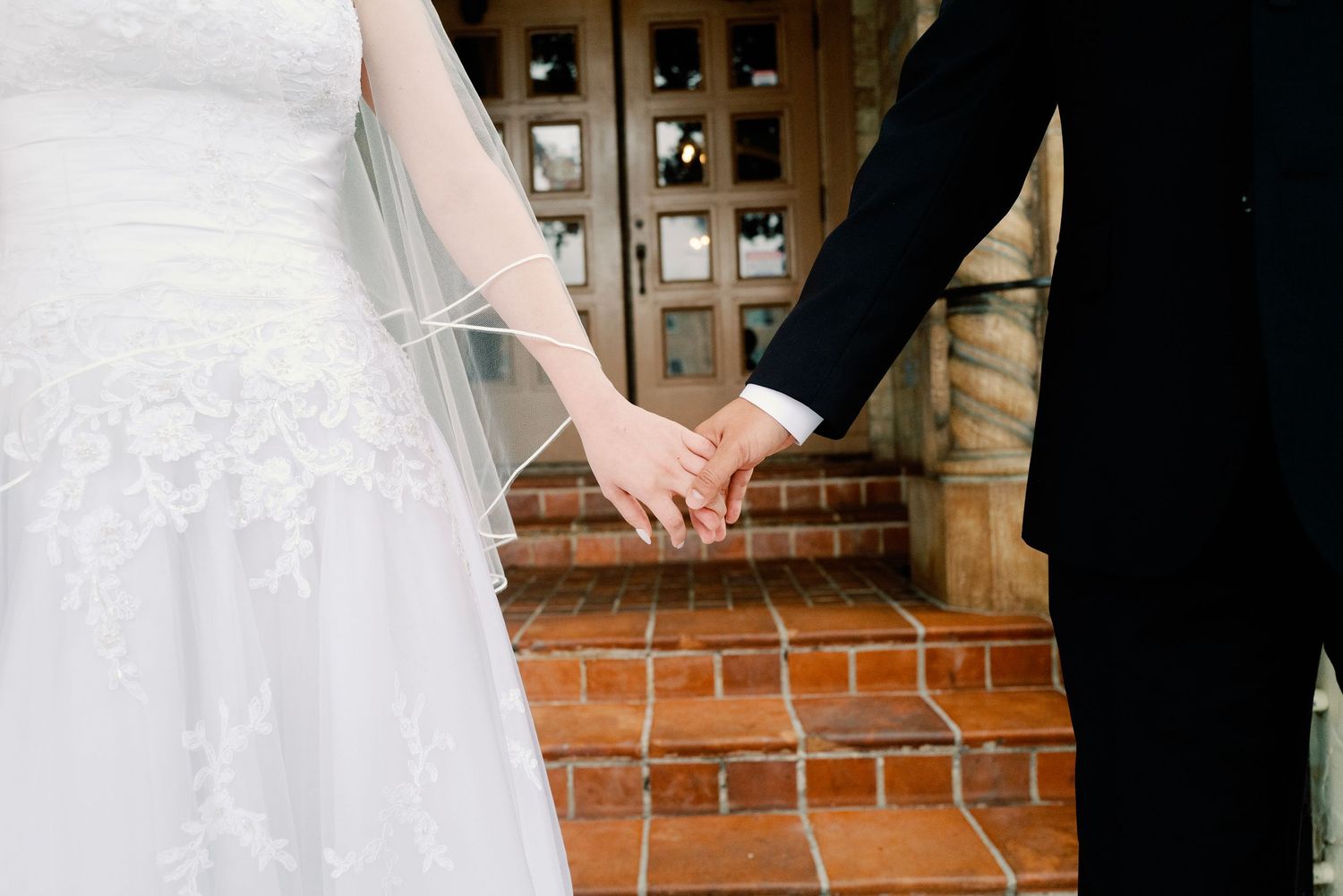 Close-up of bride and groom holding hands on brick steps in wedding attire.