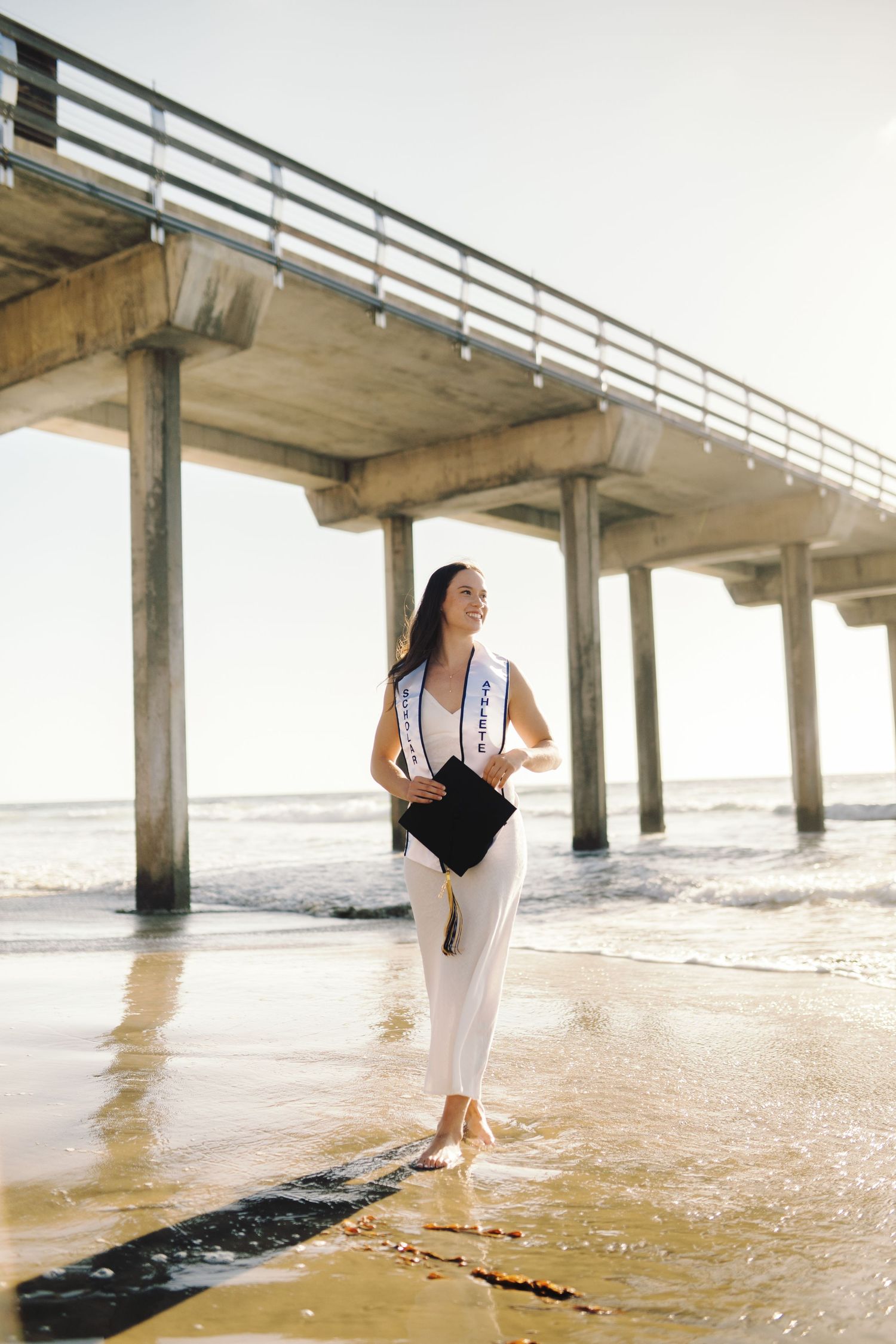 Graduate in white outfit with sash walks barefoot on beach near pier holding cap.