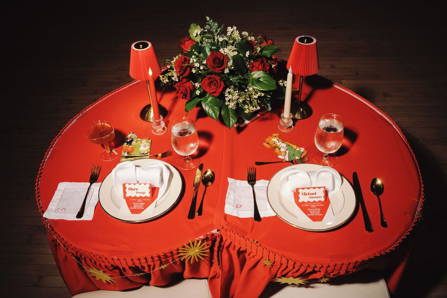 Elegant red round table set for two with gold cutlery, wine glasses, white plates, and a floral centerpiece.