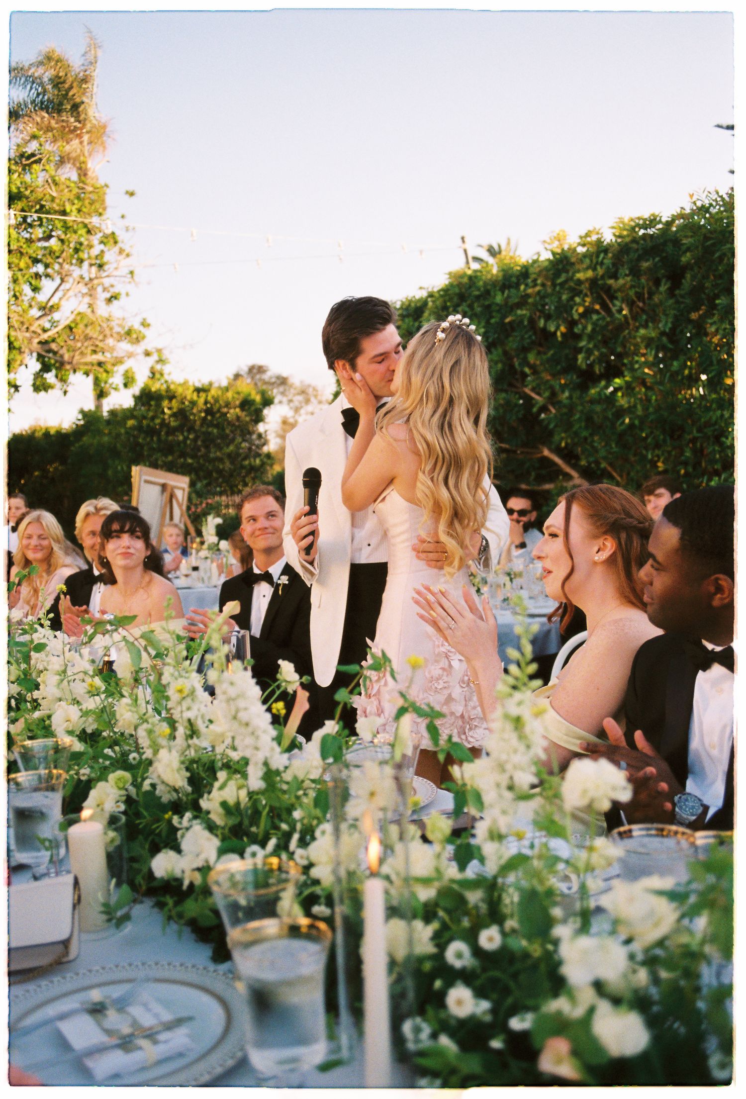 Bride and groom share a kiss at outdoor wedding reception with white floral table arrangements and guests in formal attire.