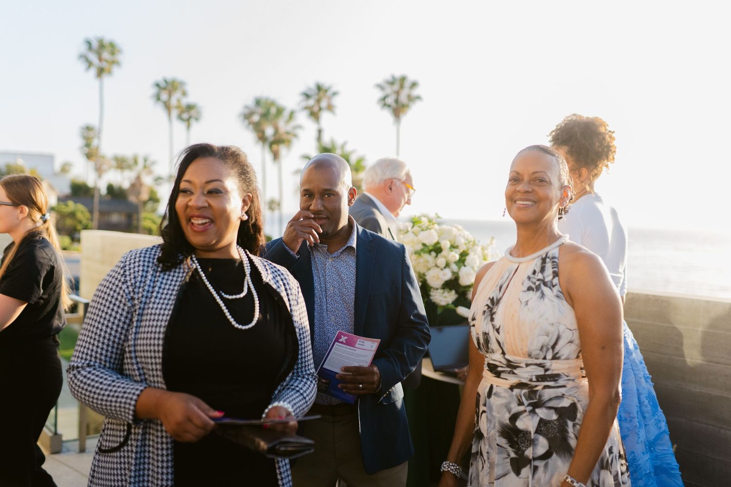 Guests smiling and socializing at an outdoor beachside event with palm trees in the background.