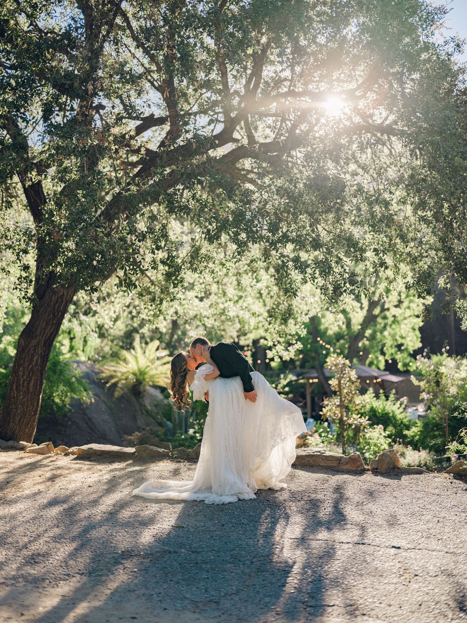Couple sharing a romantic dip kiss in a sunlit garden, bride in flowing white gown, surrounded by lush trees.