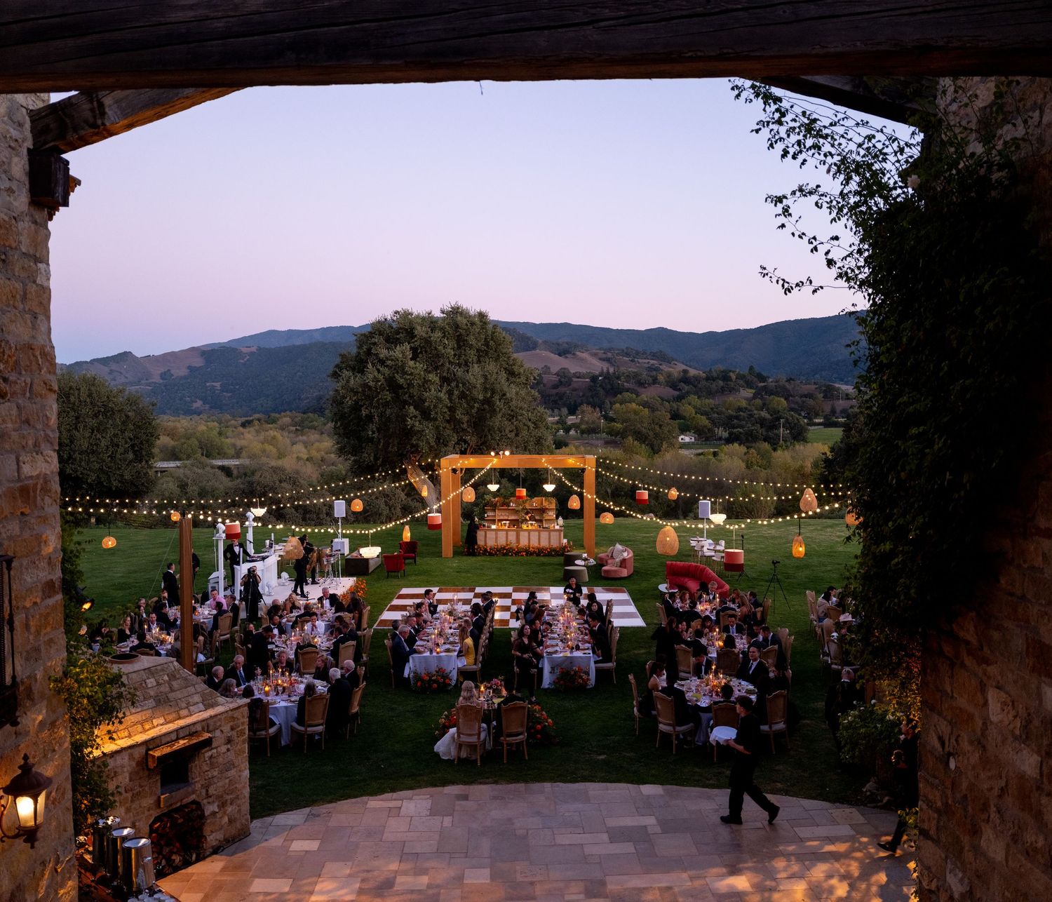 Elegant outdoor wedding reception at dusk with string lights, long dining tables, and scenic mountain views.