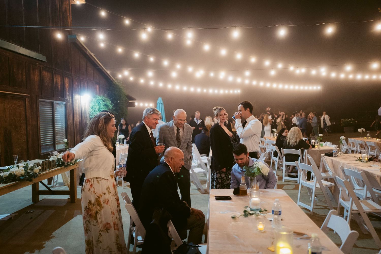 Wedding reception guests mingling under string lights inside a rustic barn venue.