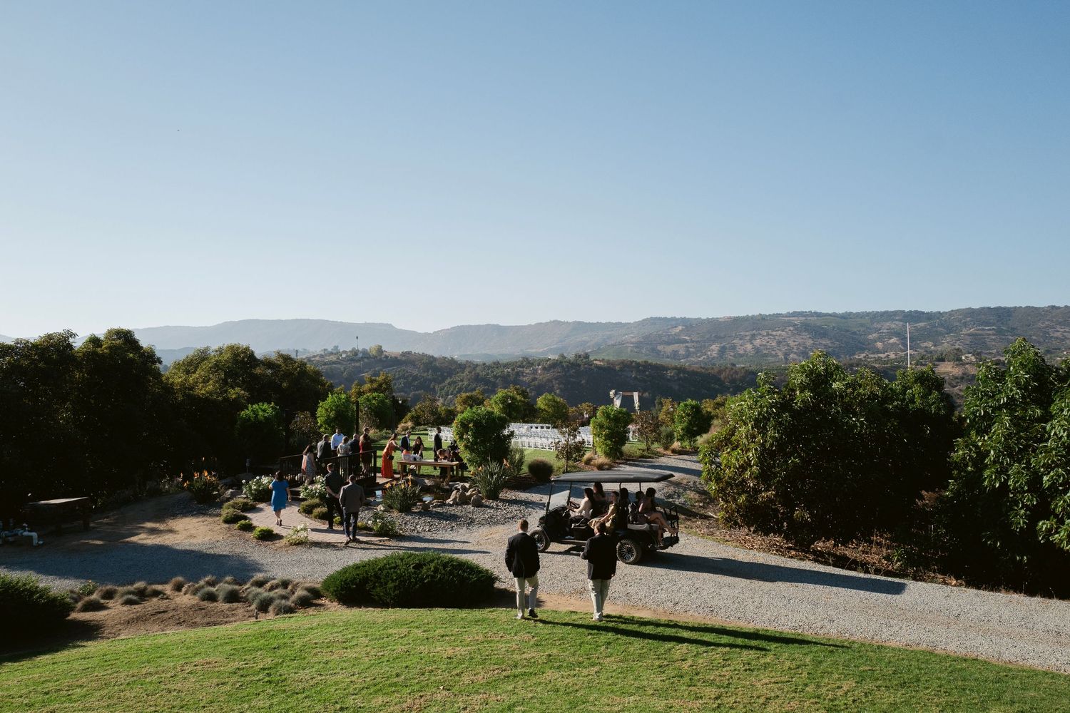 Wedding guests walk through scenic outdoor venue with rolling hills and blue sky backdrop.