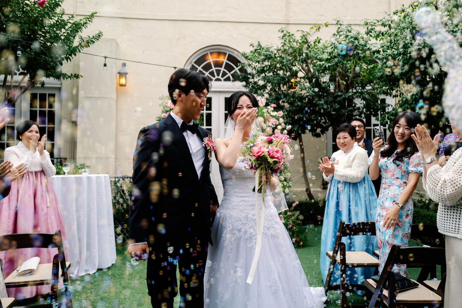 Bride and groom exit ceremony through bubble shower as guests celebrate at outdoor wedding.