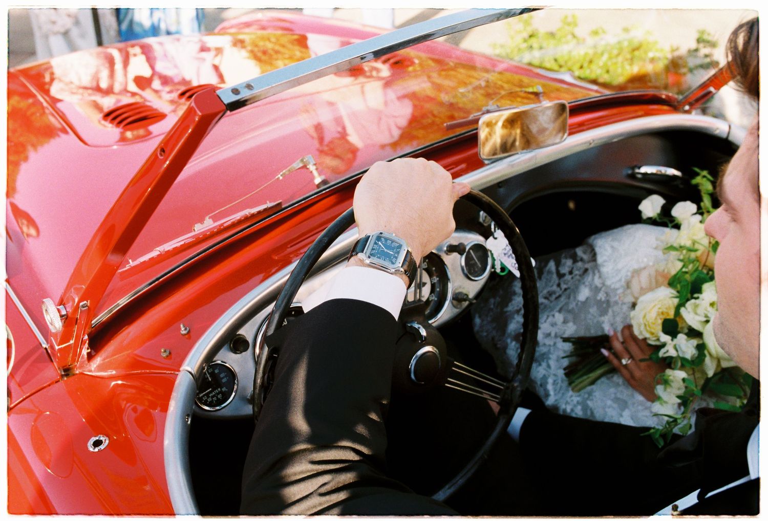 Groom drives classic red convertible with bridal bouquet resting on dashboard after wedding ceremony.