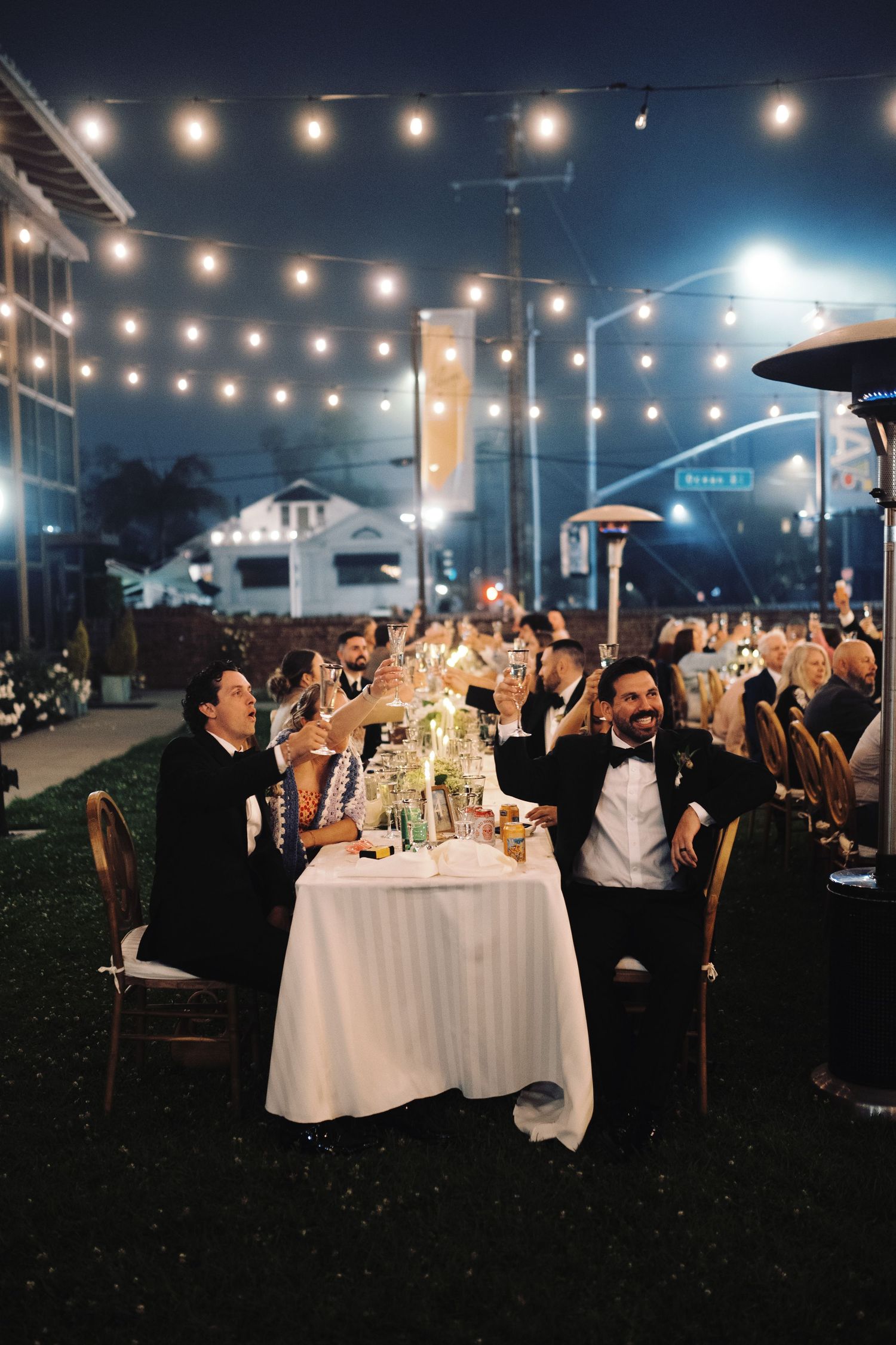 Wedding party raises glasses in a toast at outdoor night reception under string lights and patio heaters.