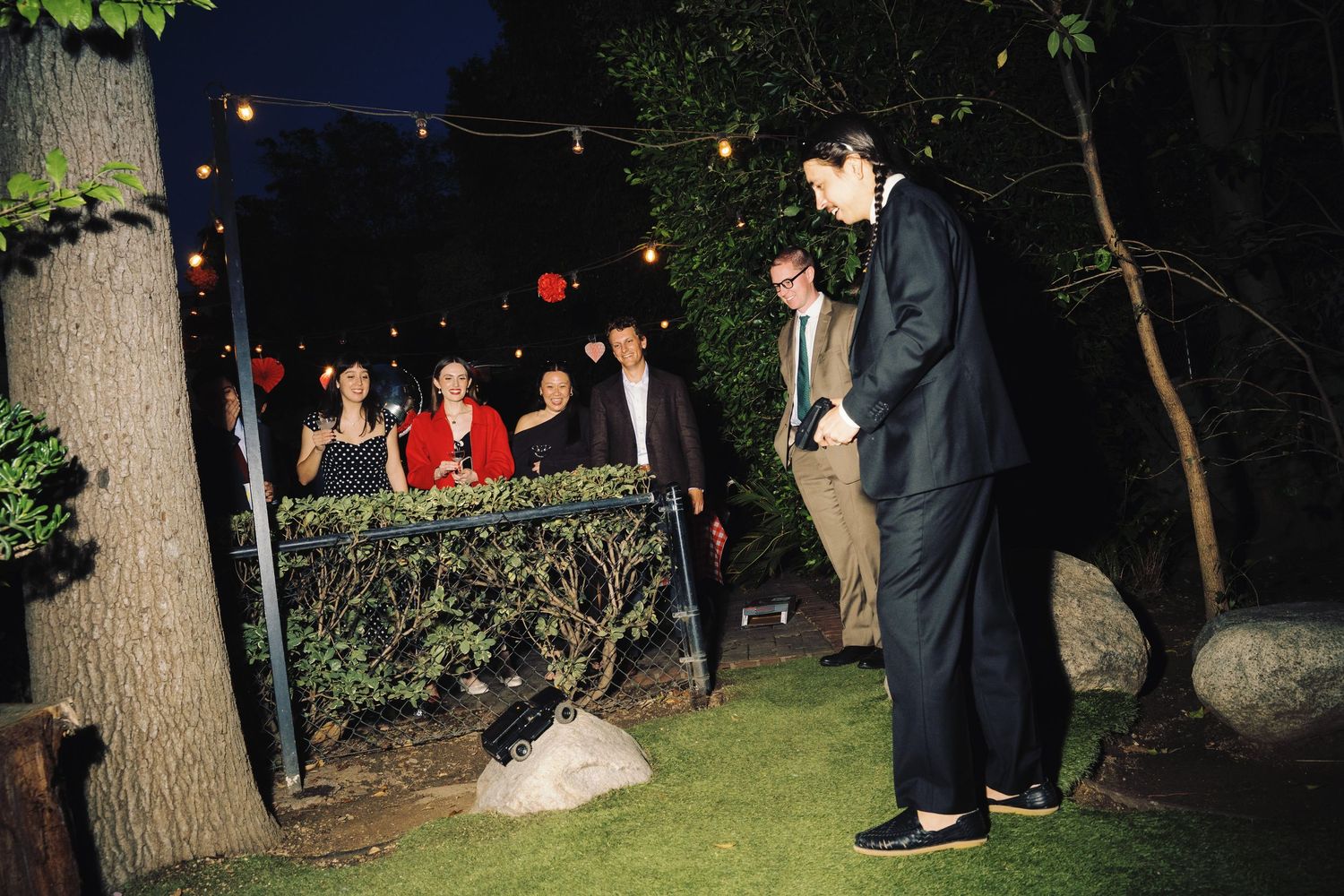 Guests socializing outdoors at a nighttime garden party with string lights and trees in the background.