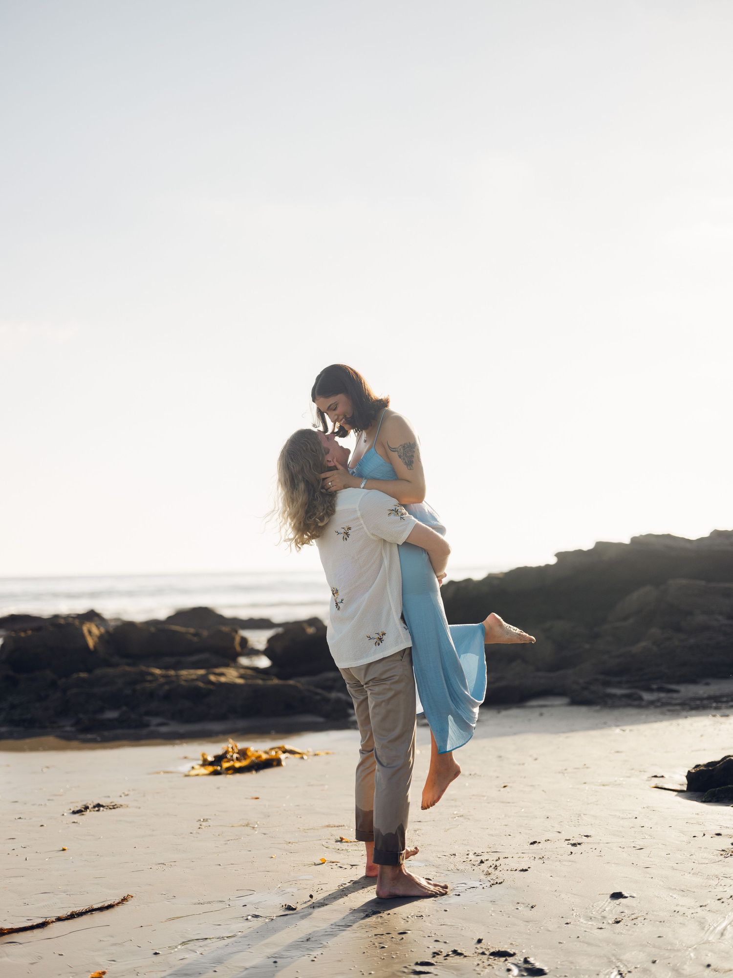 Couple sharing a kiss on sandy beach at golden hour, man lifting woman in flowing blue dress near rocky cliffs.