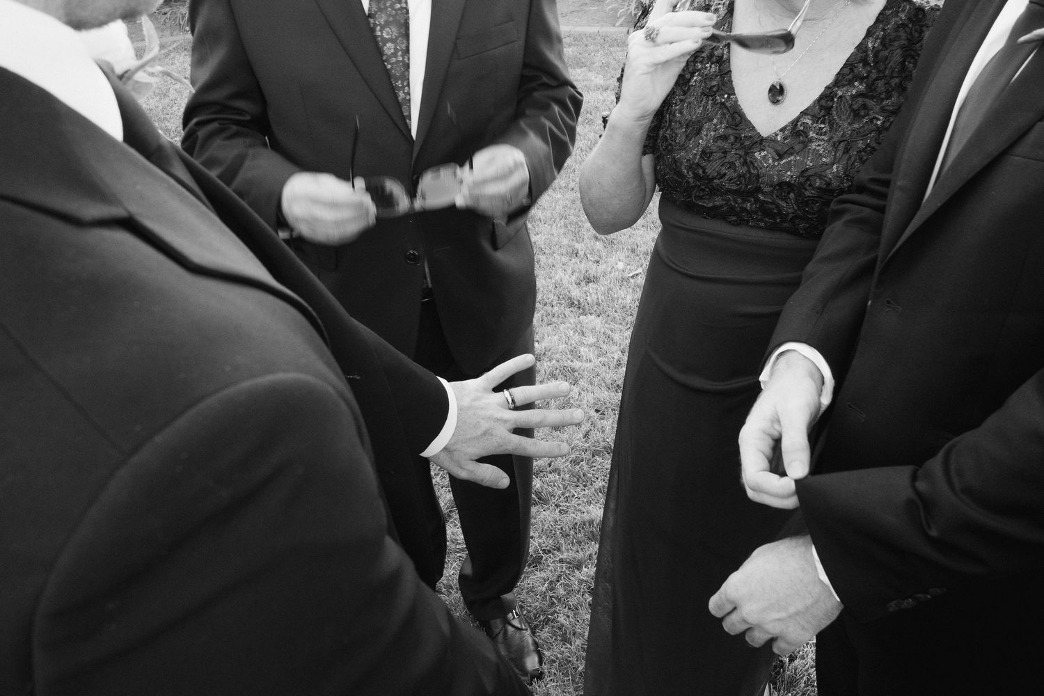 Black and white close-up of wedding guests in formal attire conversing and gesturing on a grassy lawn.