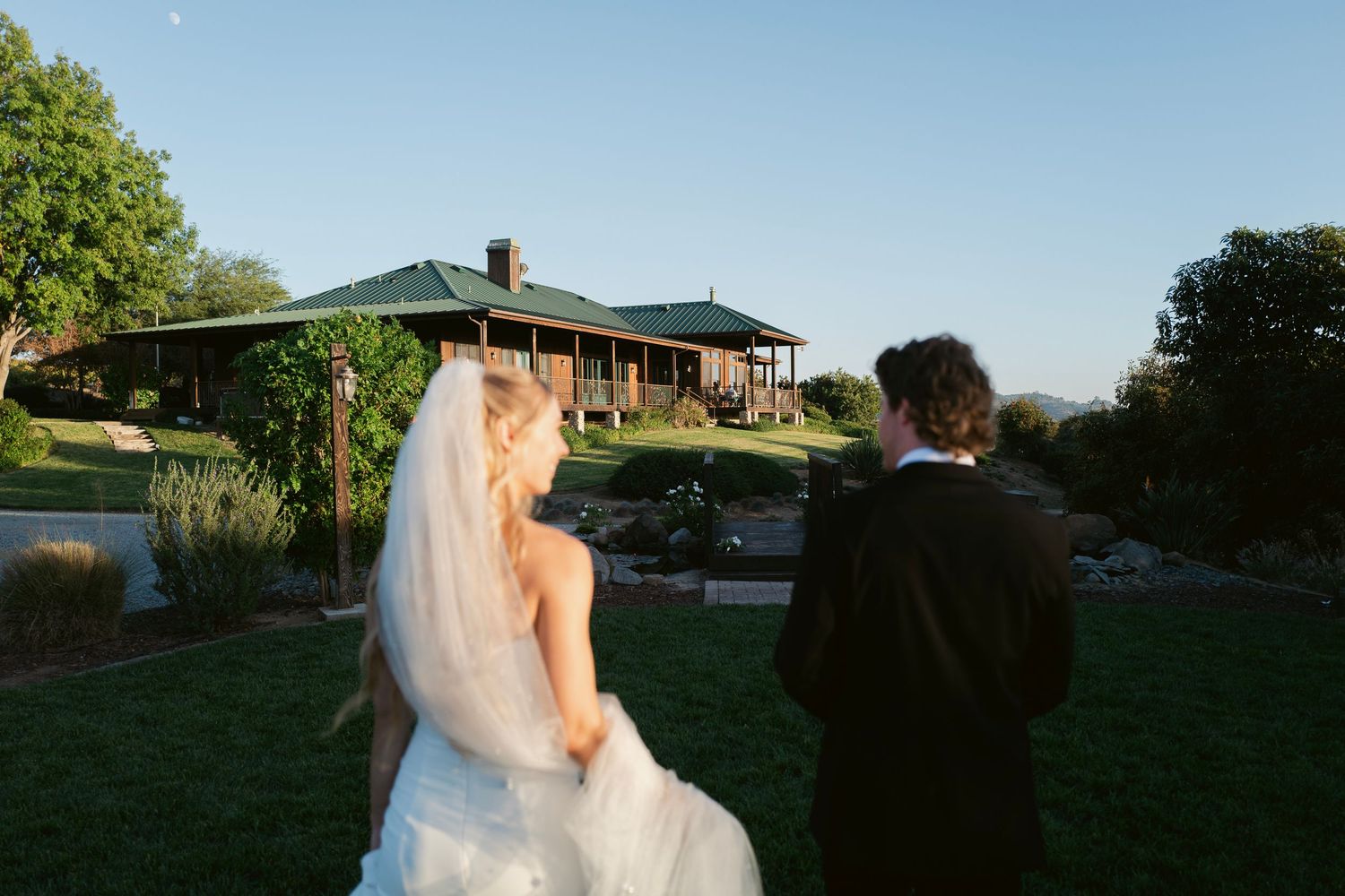 Bride and groom walk hand in hand toward a rustic venue surrounded by lush gardens at golden hour.