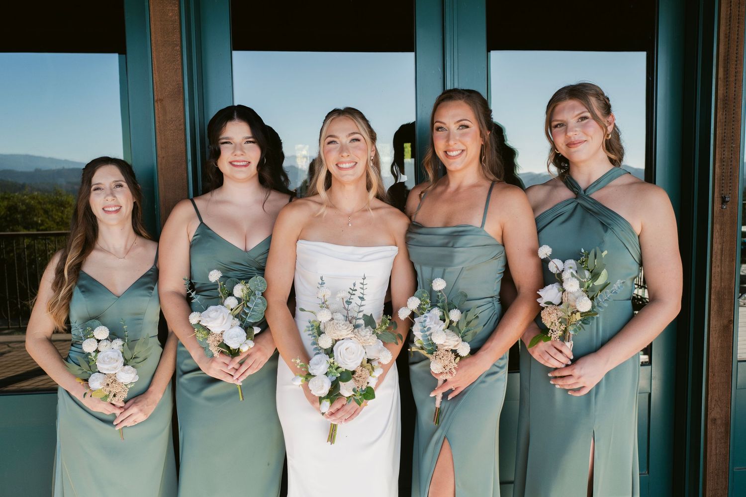 Bride with four bridesmaids in sage green dresses holding white bouquets at scenic venue.