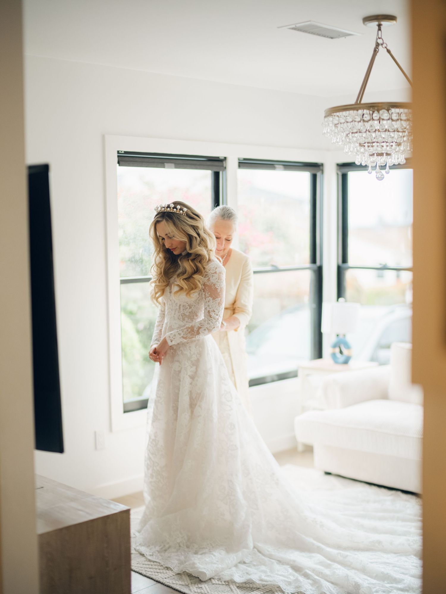 Bride in elegant long-sleeve lace wedding gown with tiara being helped by older woman in bright modern room.