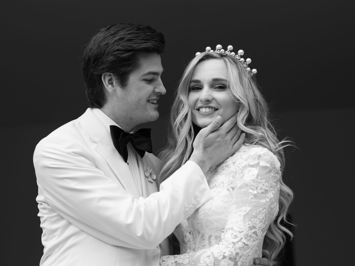 Black and white photo of groom tenderly holding bride's face as she smiles wearing a pearl crown.