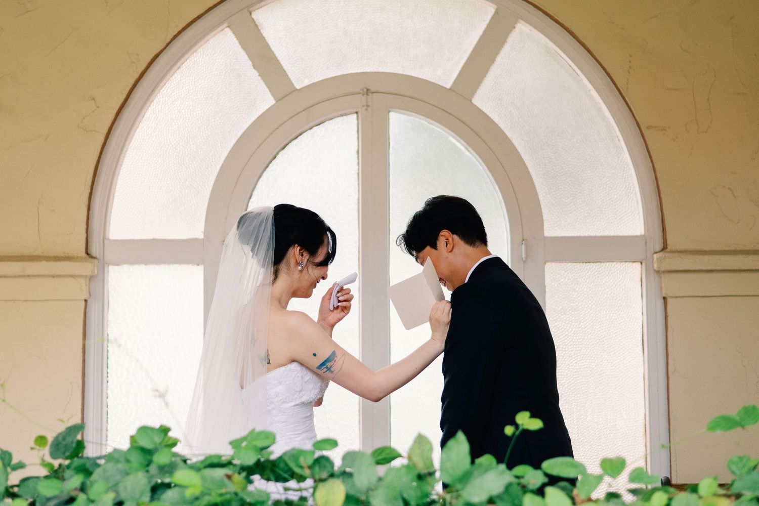 Bride and groom sharing tender moment exchanging gifts by arched window.