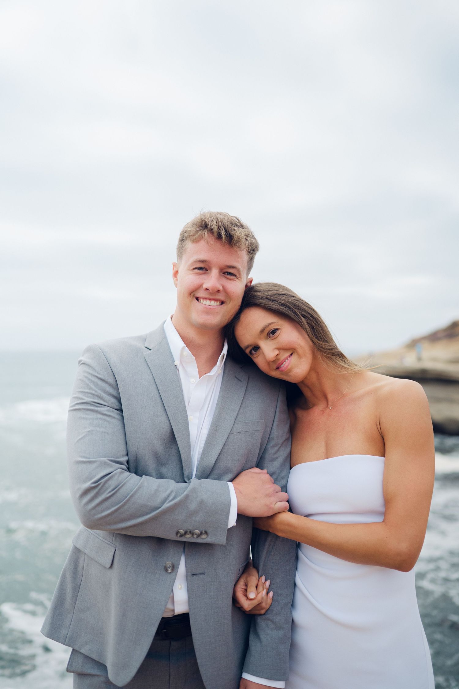 Smiling engaged couple posing together in formal attire on a rocky oceanside cliff.